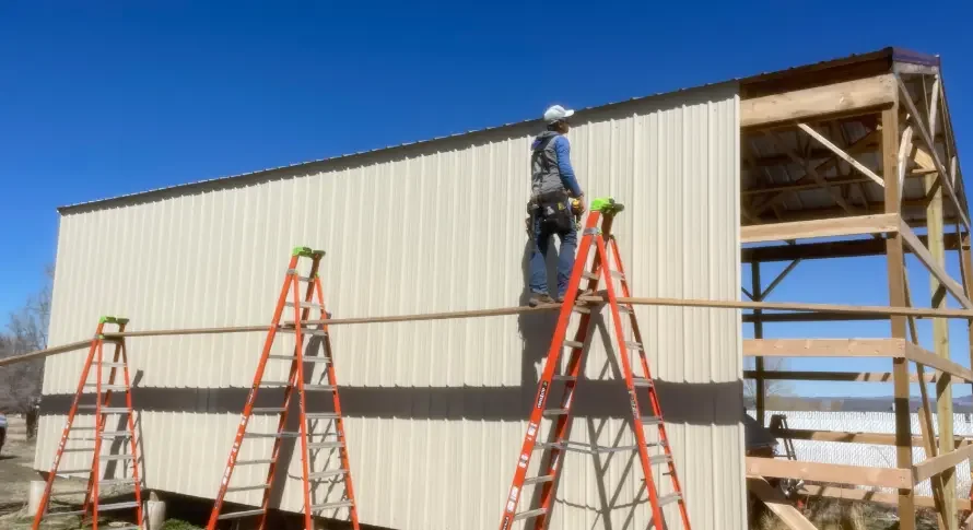 Construction worker standing on a ladder, working on the exterior wall of a building under construction, with a wooden framework visible.