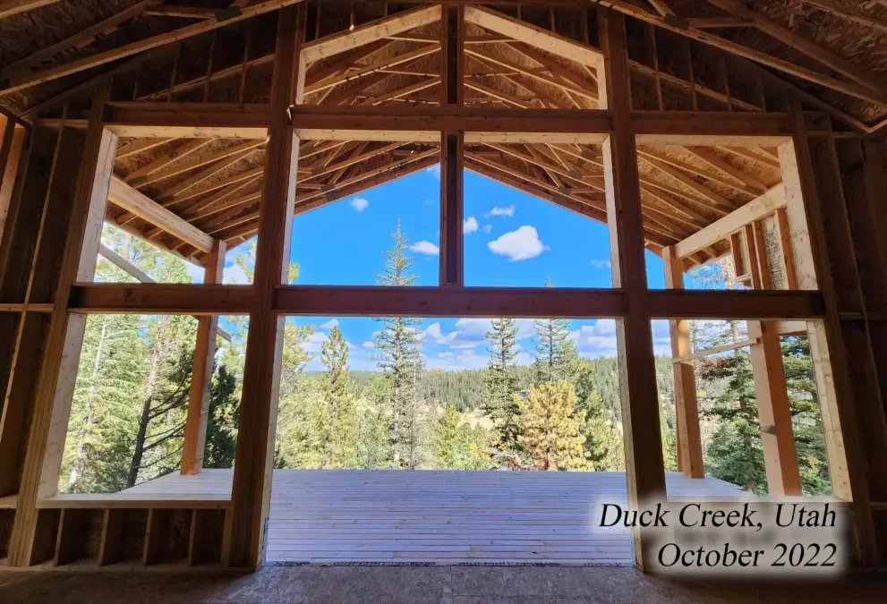 View of a lush, green forest with tall trees seen through the unfinished wooden frame of a building under construction, with a clear blue sky and a few clouds. Text in the bottom right corner reads: "Duck Creek, Utah October 2022."