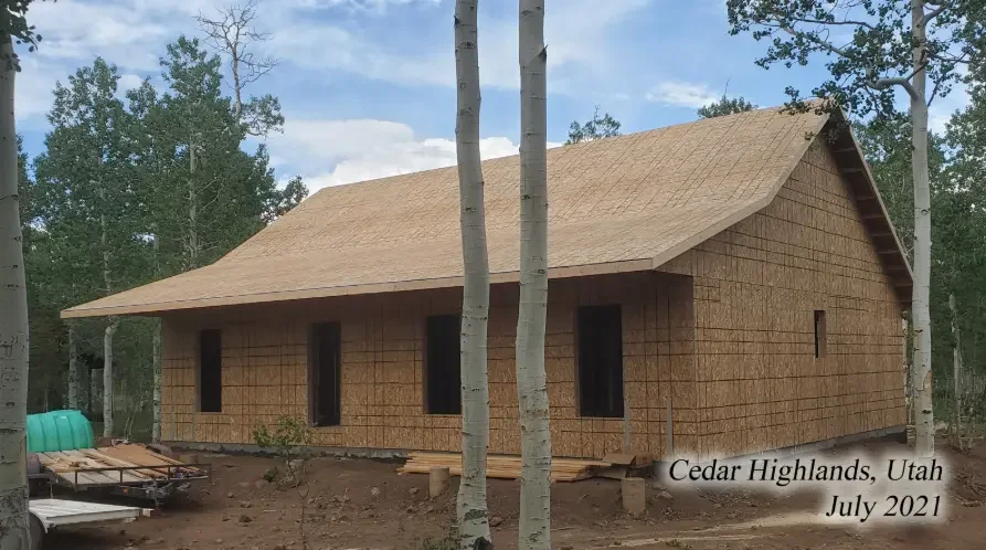 A wooden house under construction in Cedar Highlands, Utah, July 2021, surrounded by trees with a partly cloudy sky above.