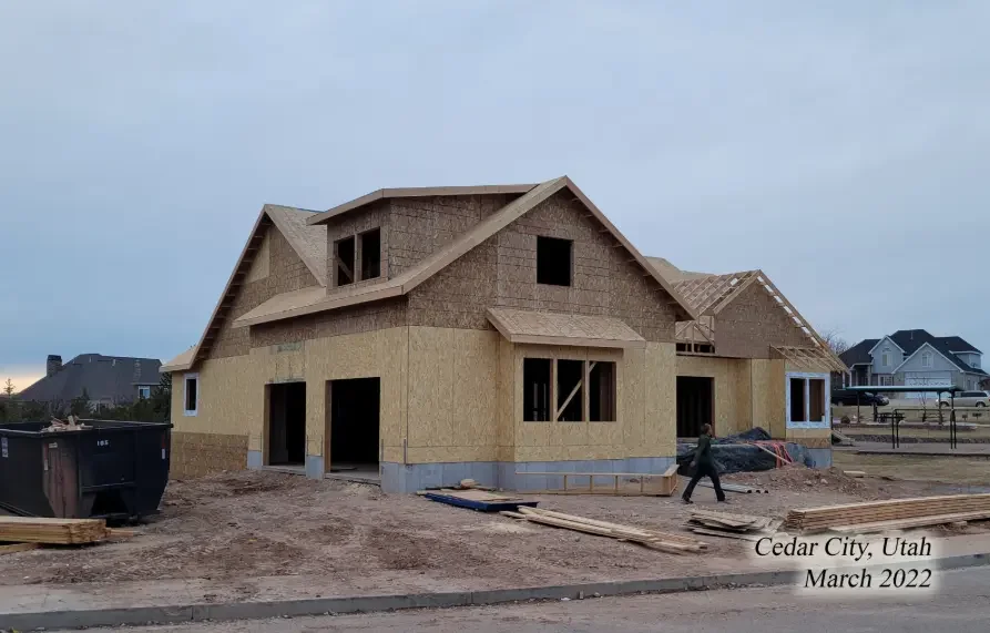 Under construction house in Cedar City, Utah, with wooden framing and sheathing, and a person walking nearby, March 2022.