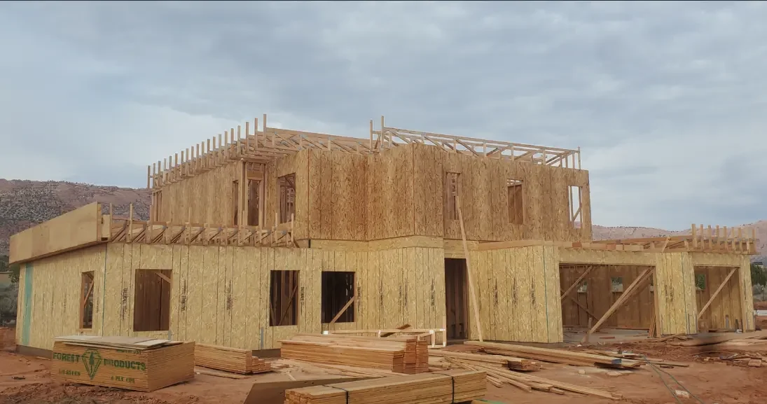 A two-story house under construction with wooden framing and plywood sheathing, set in a semi-arid landscape under a cloudy sky.