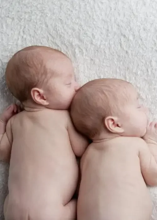 Two sleeping infants lying on a soft surface, side by side, with their heads touching and faces turned to the side.