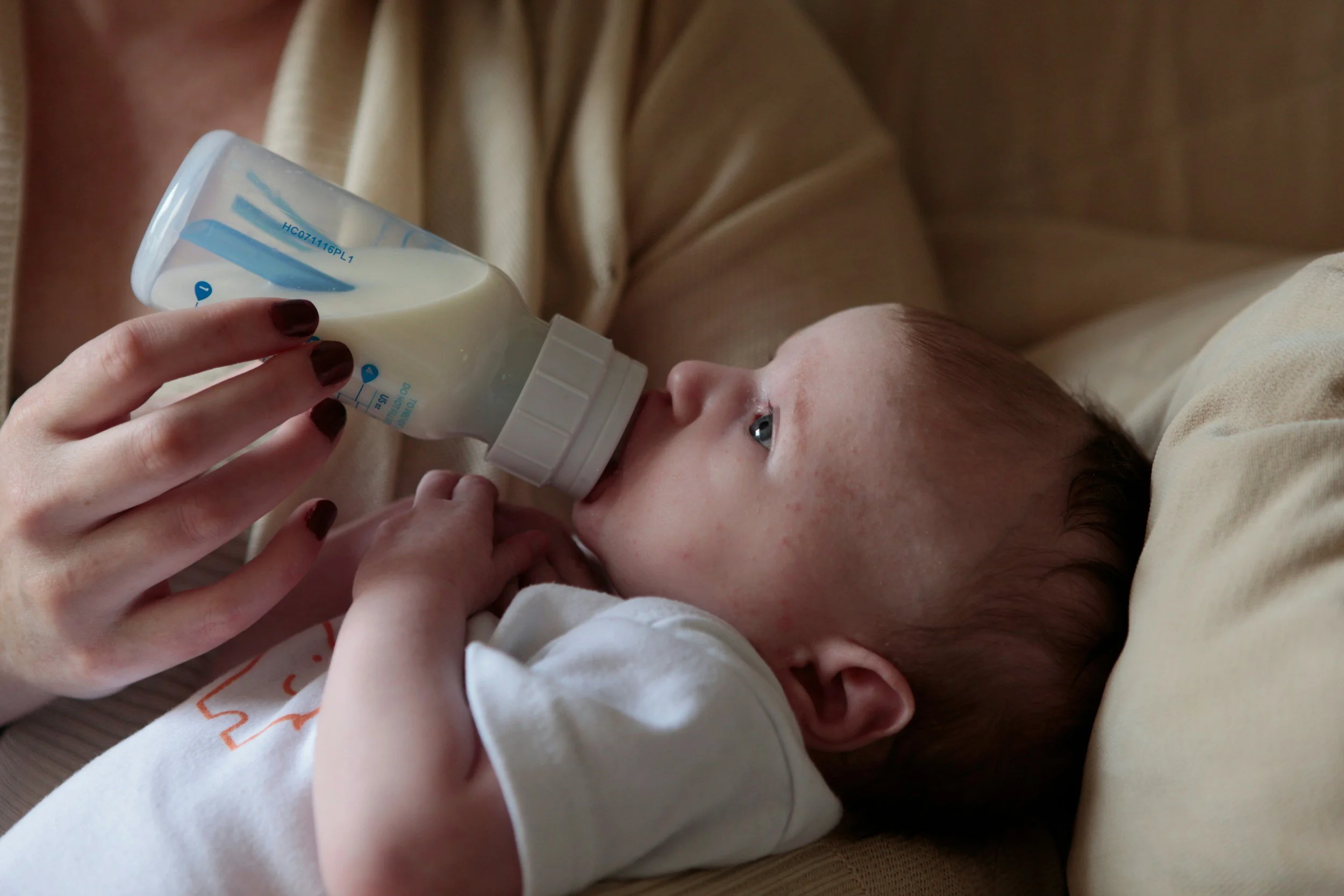A young child with brown hair and blue eyes lying on a beige cushion, drinking from a baby bottle that a hand is holding for them.