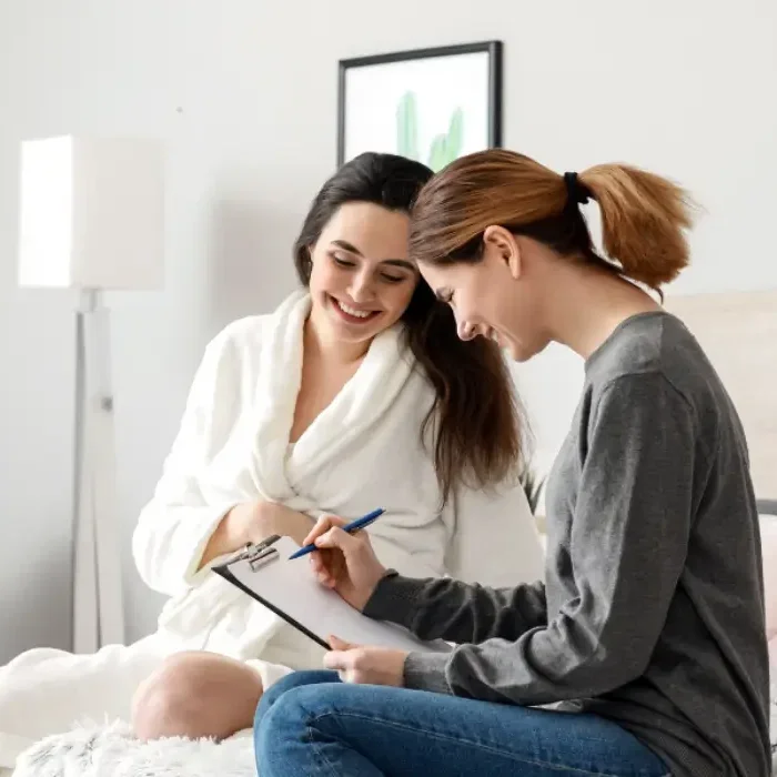 A woman in a gray shirt filling out paperwork on a clipboard while talking to a woman in a white bathrobe. They are sitting on a bed in a bright, modern room.