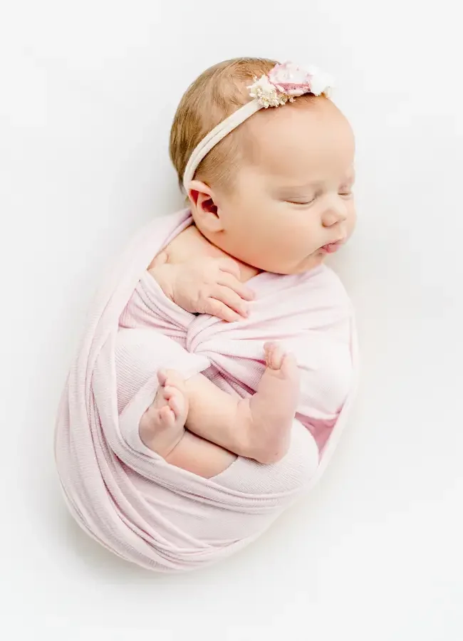 A newborn baby girl sleeping, wrapped in a light pink blanket with a pink floral headband.