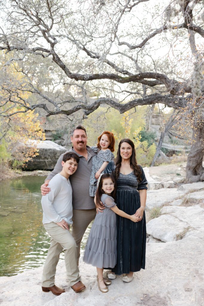 A family of five standing on a riverbank outdoors under a large, leafless tree with rocks and greenery in the background. The father and the daughter are smiling at the camera.