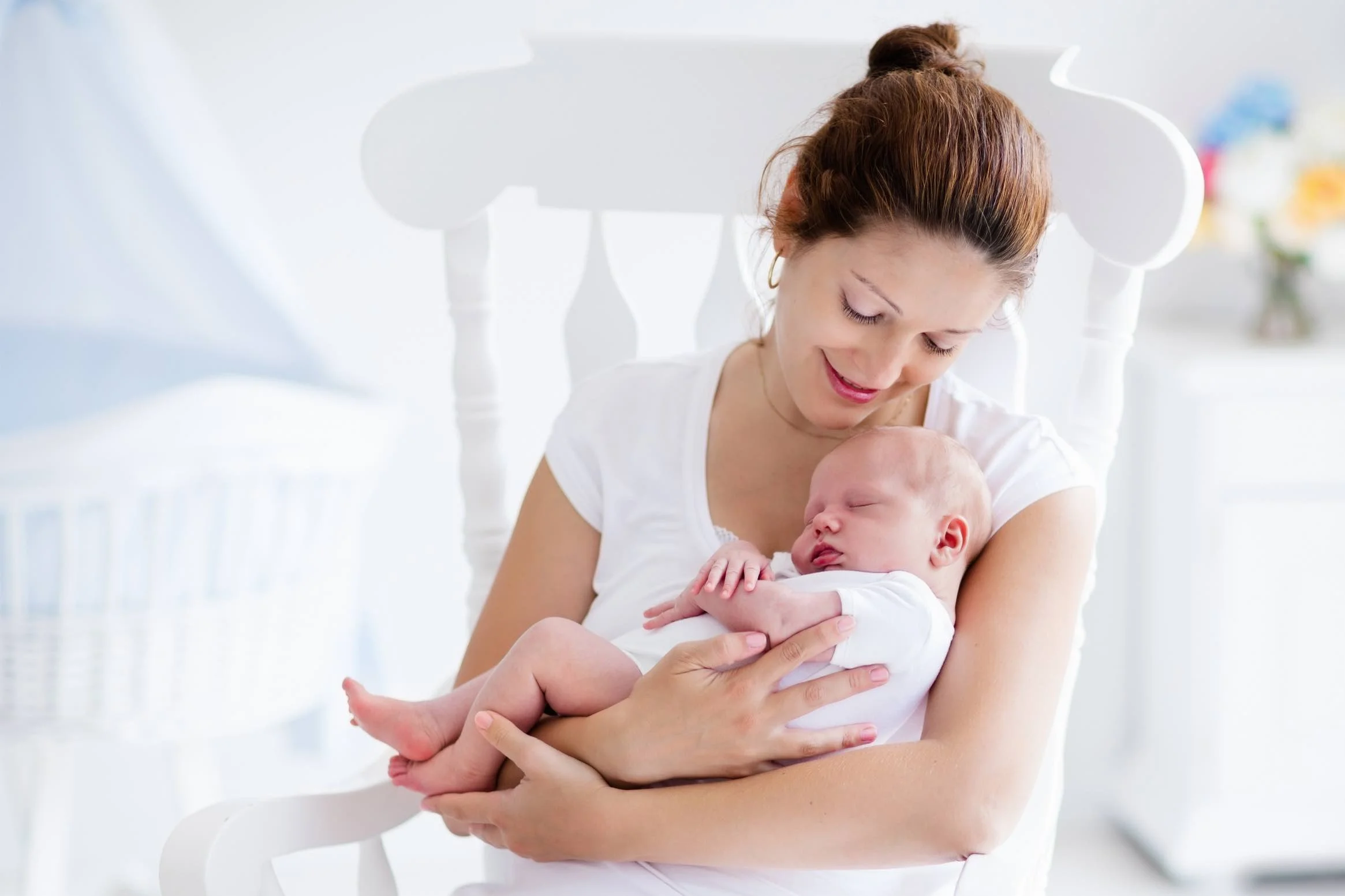 A woman with reddish hair tied up in a bun, smiling and looking down, holding a sleeping baby in her arms in a bright, white room.