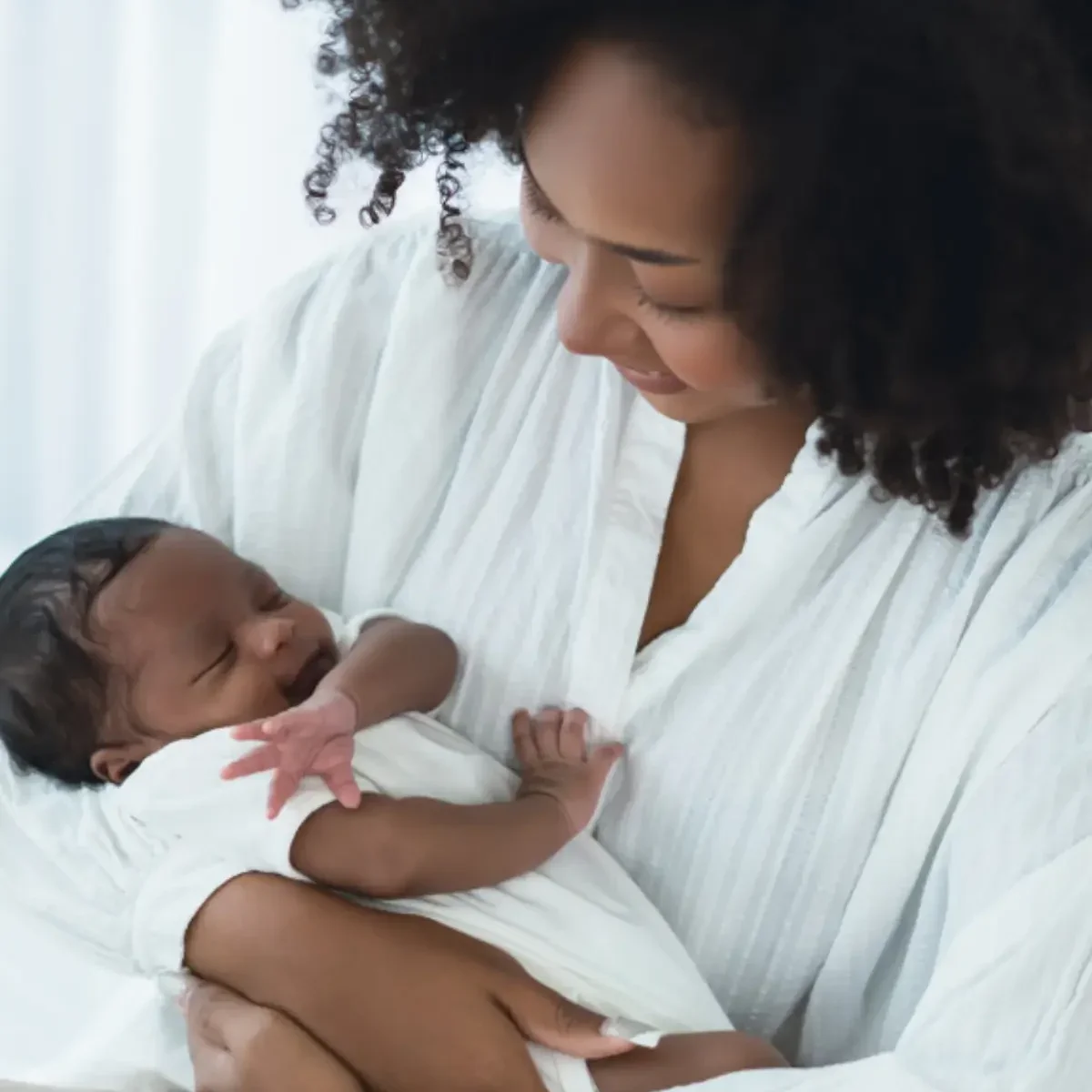 A woman with curly hair holding a newborn baby in a white hospital gown, both smiling and looking at each other.