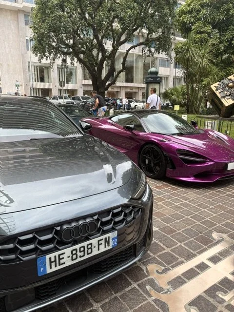 Deux voitures de sport, une noire et une violette, stationnent sur une place pavée à côté d'un arbre.