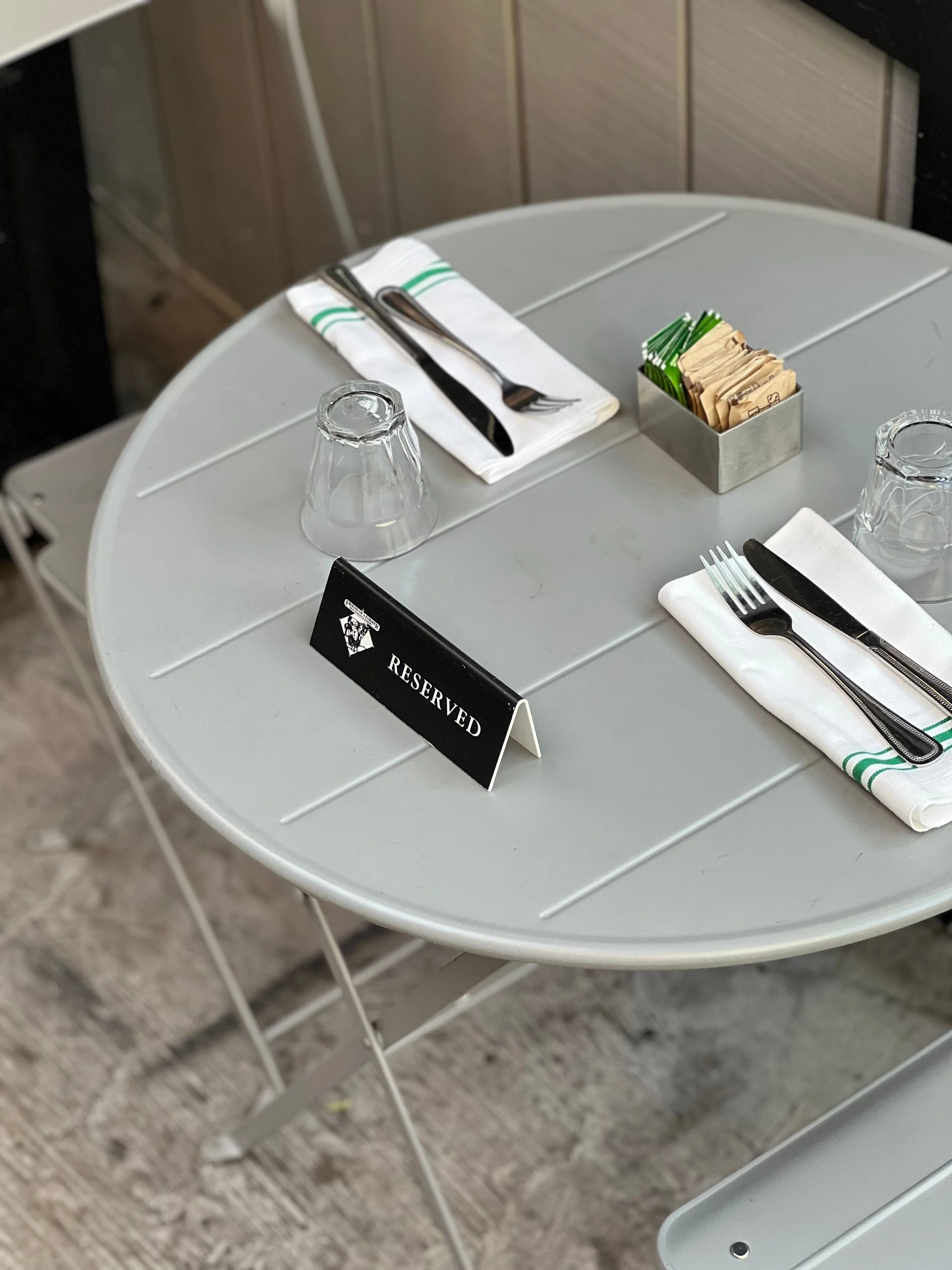Une table réservée à une clientèle, avec un signe 'Réservé', des couverts, des verres, et des sachets de sucre ou de crème dans un récipient en métal.