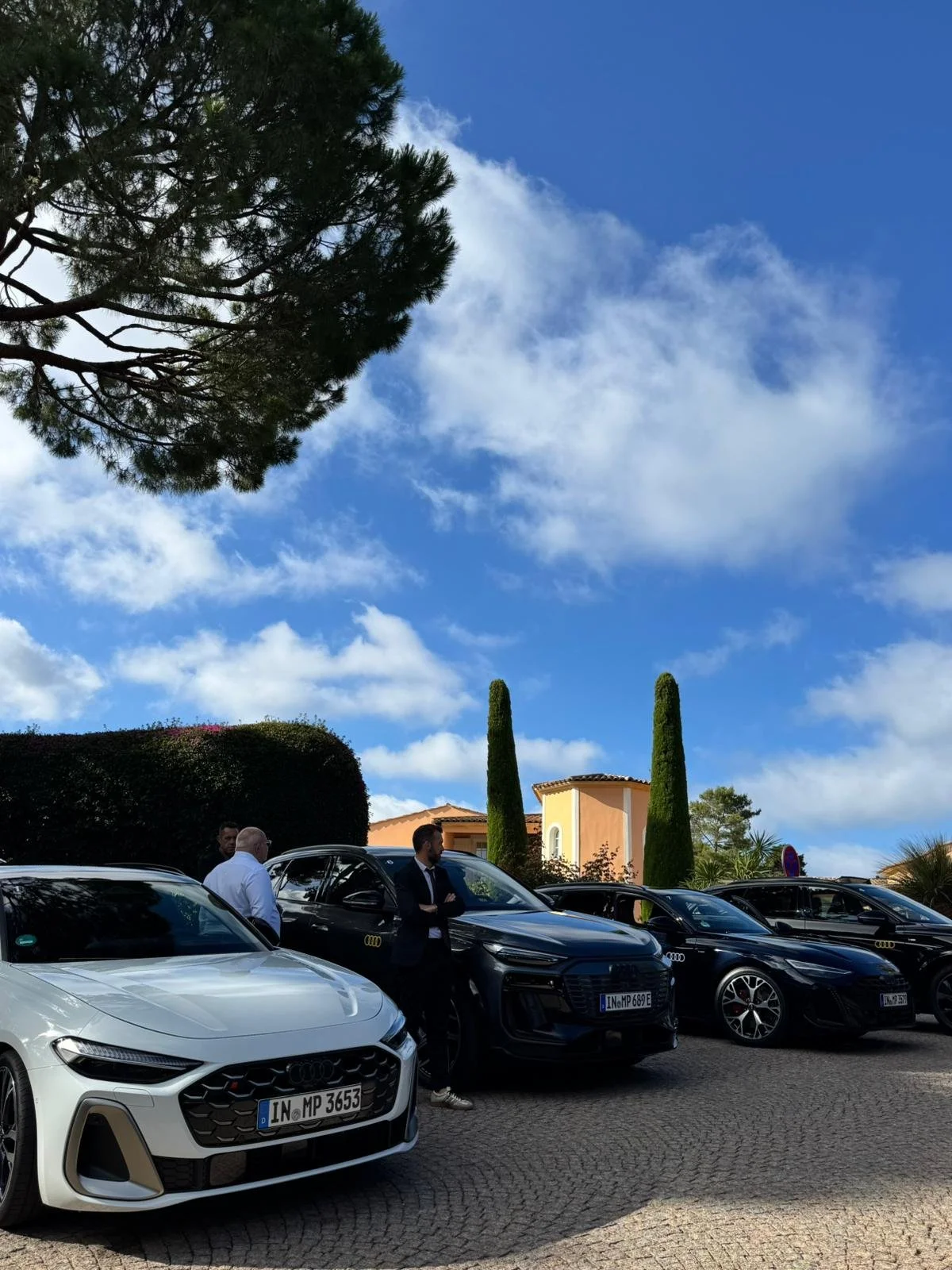 voiture Audi blanche et huit voitures noires garées devant une maison ensoleillée avec des arbres verts et un ciel bleu avec des nuages