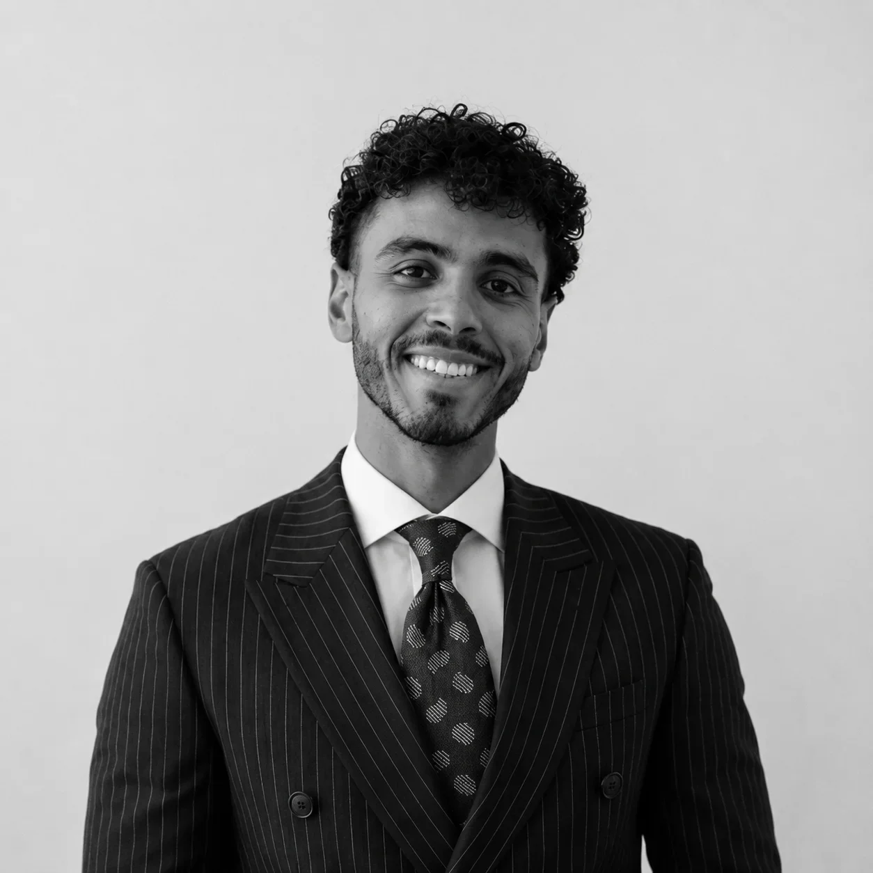 Jeune homme souriant en costume rayé, chemise blanche et cravate à motifs, portrait en noir et blanc, fond neutre.