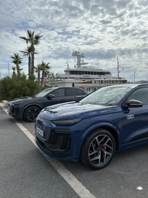 Deux voitures Audi, une bleue et une noire, garées dans une rue près d'un port avec un grand yacht en arrière-plan et des palmiers, sous un ciel partiellement nuageux.