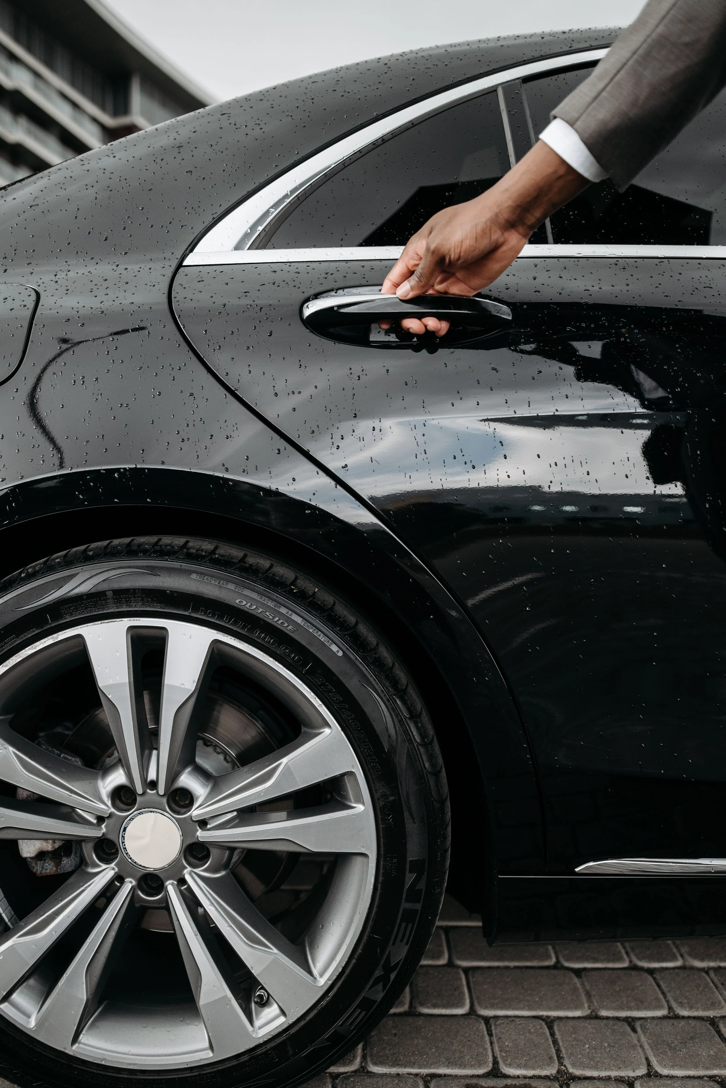 Une main qui ouvre la porte d'une voiture noire en plein jour, la voiture est mouillée de pluie, avec une jante en aluminium visible et un fond pavé.