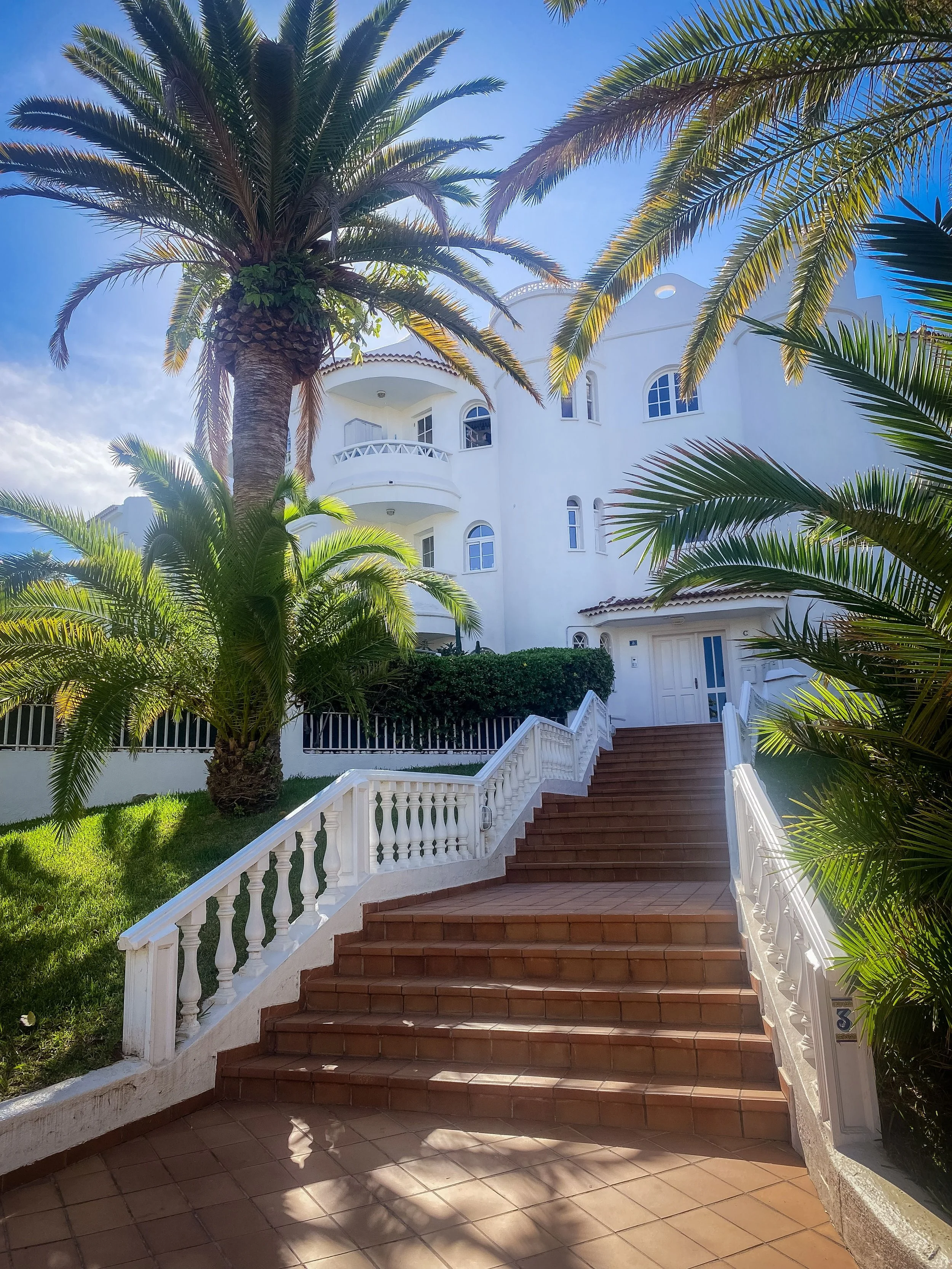 Escalier menant à une maison blanche avec balcon, entourée de palmiers, sous un ciel ensoleillé.