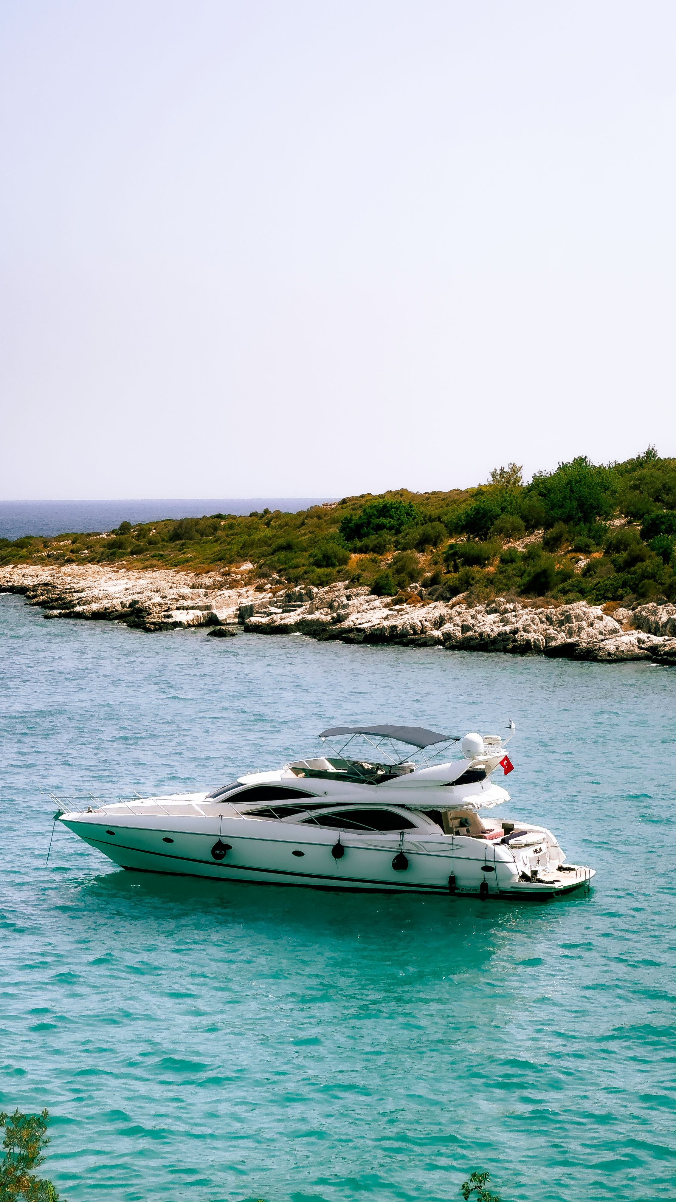 Un yacht blanc flottant sur une mer calme avec une côte rocheuse et des arbustes verts en arrière-plan.