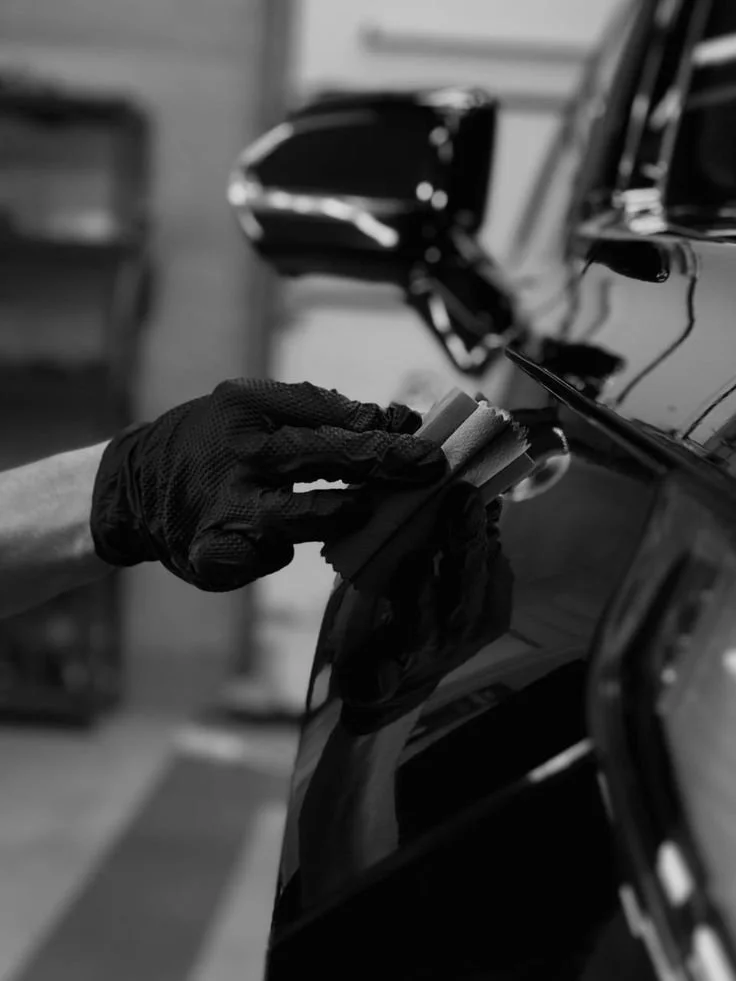 Keramikversiegelung Hände in schwarzen Handschuhen, die einen Lackierpinsel halten, neben einem schwarzen Auto in einer Werkstatt.