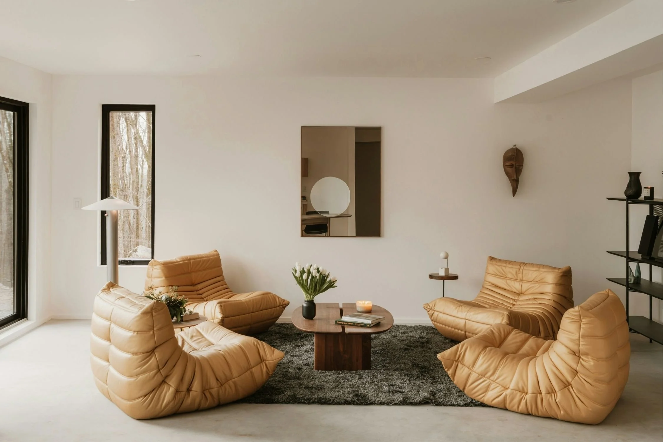 Modern living room with tan lounge chairs, round coffee table, gray rug, wall art, and tall windows letting in natural light.