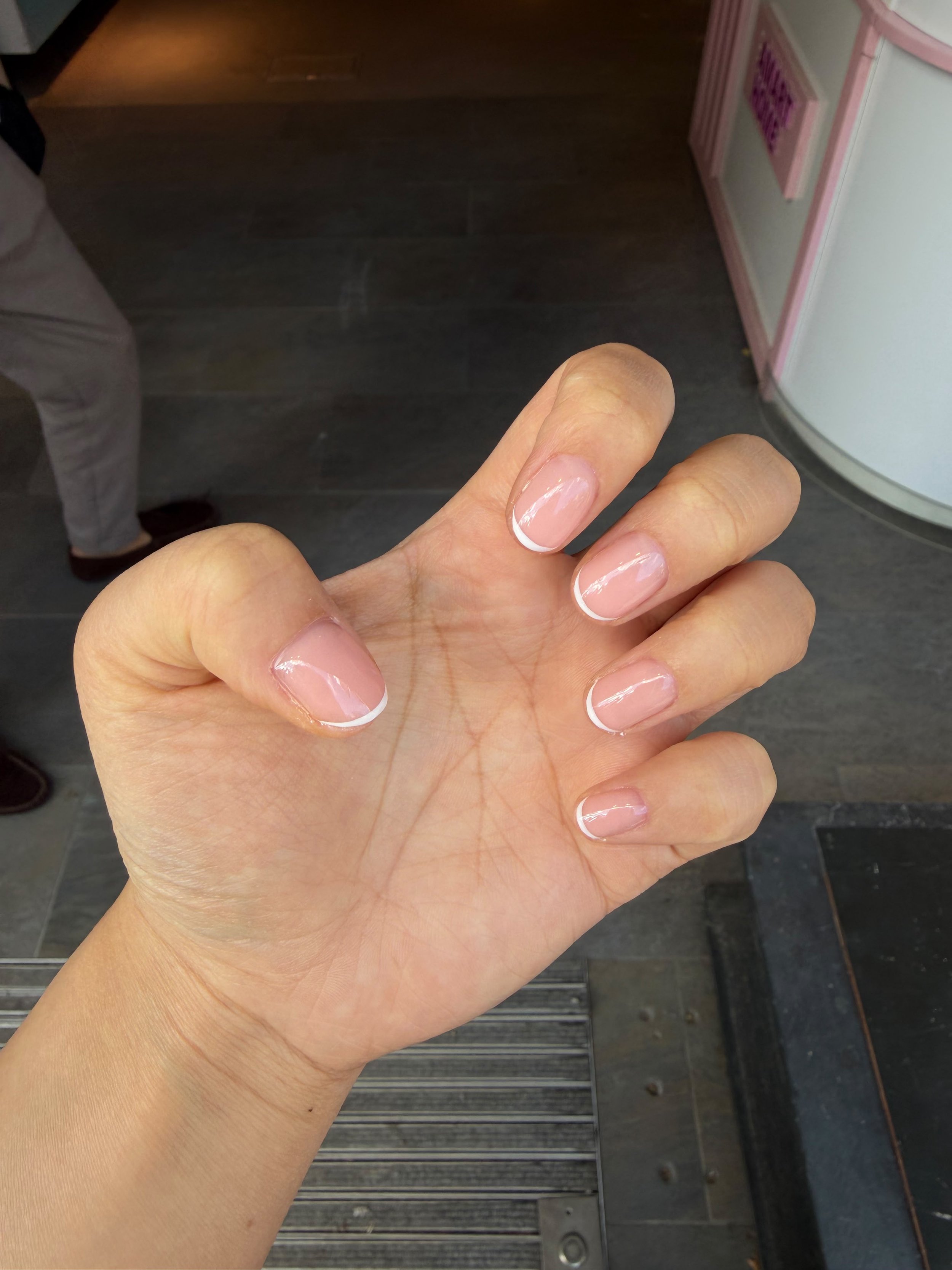 Close-up of a hand with French manicure nails in front of a dark floor and a pink and white wall or counter.