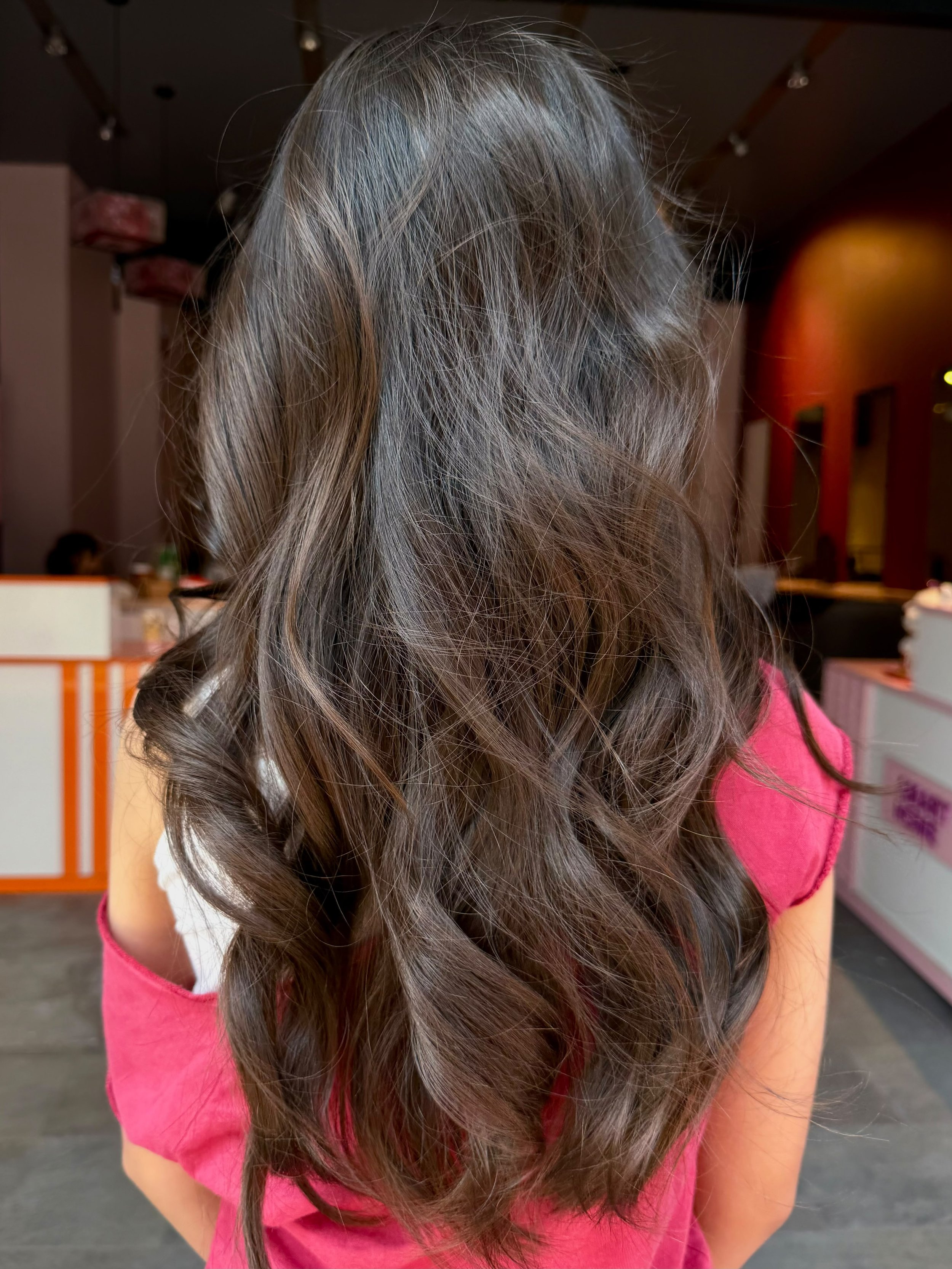 Close-up of a person's head with long, wavy, brown hair, wearing a pink shirt, in an indoor setting with colorful lights and furniture in the background.
