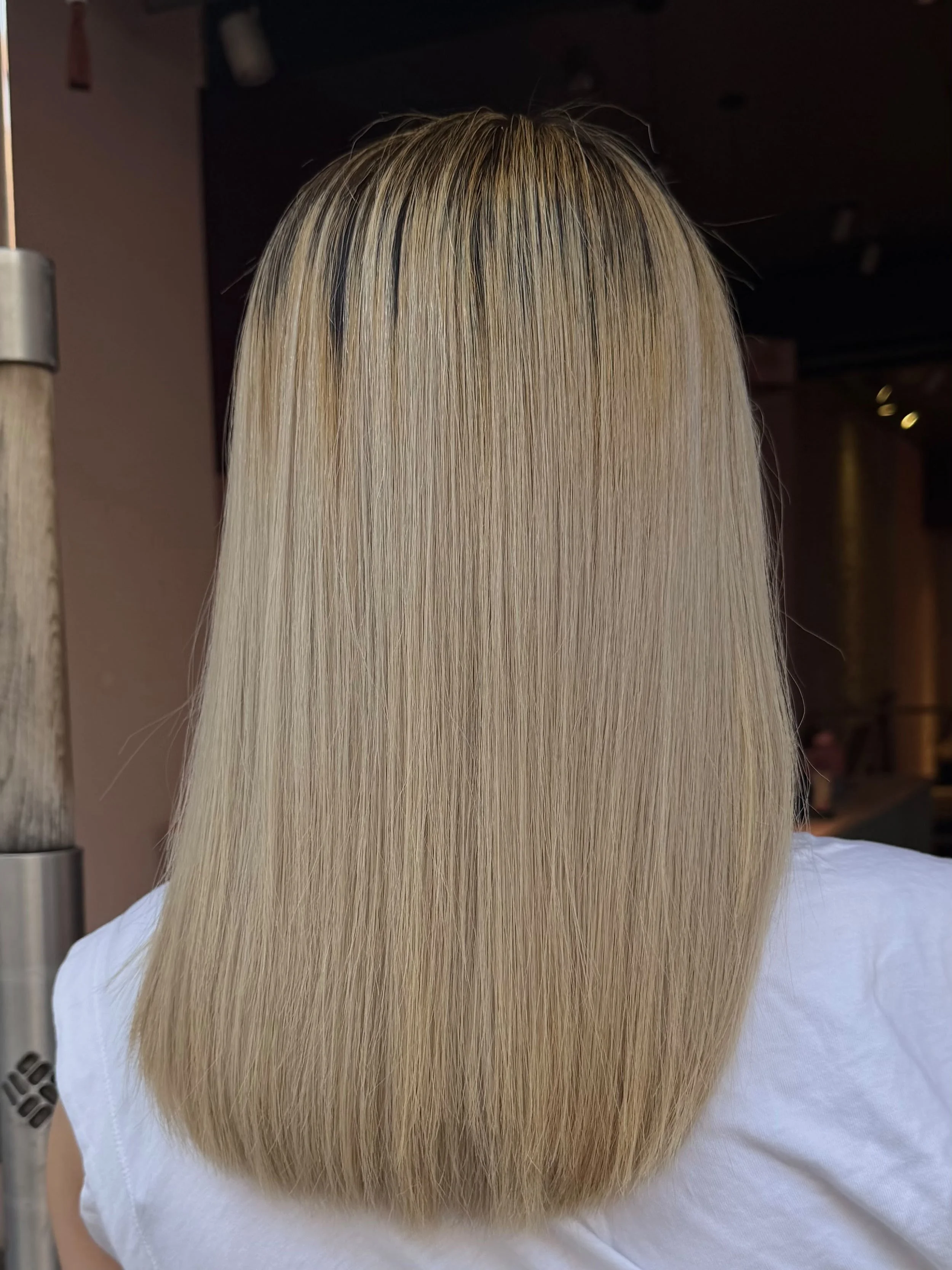 Back view of a person with long, straight, blonde hair, wearing a white shirt, in a salon or similar setting.