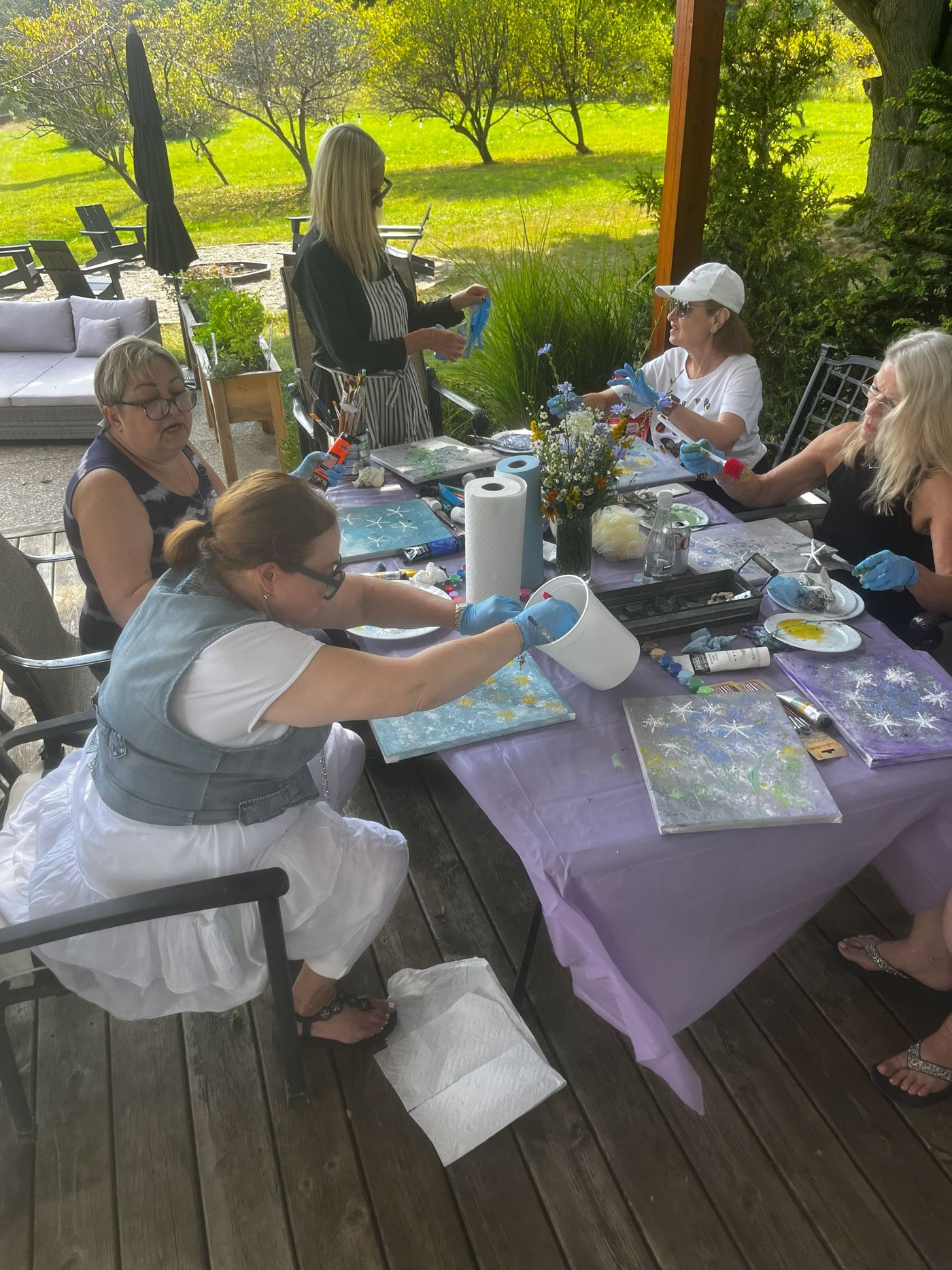 People participating in a paint and sip class outdoors on a covered patio, with paintings and painting supplies on the table, surrounded by a lush green backyard.