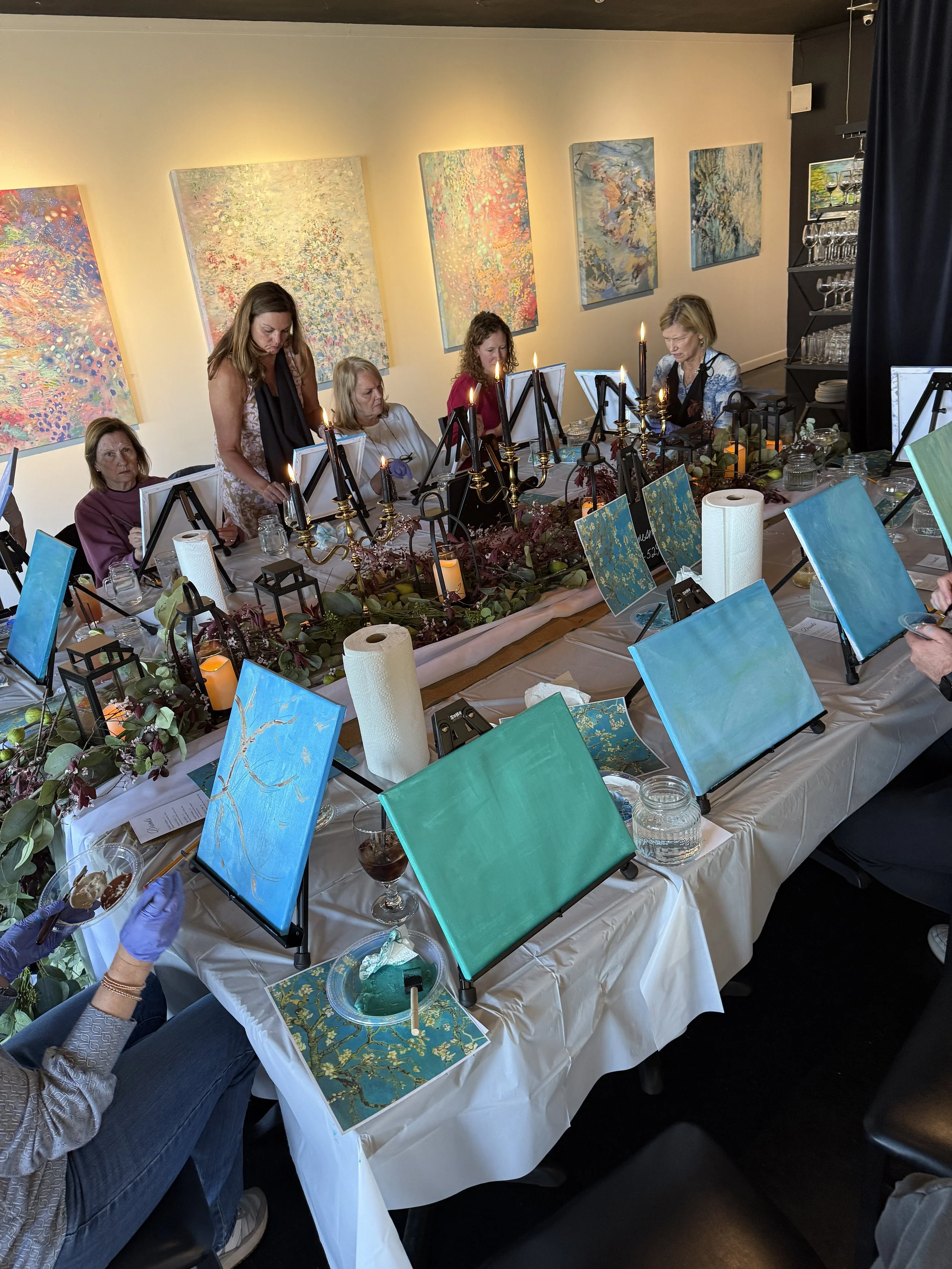 People painting on canvases arranged on a long table in an art studio with colorful abstract paintings on the wall.