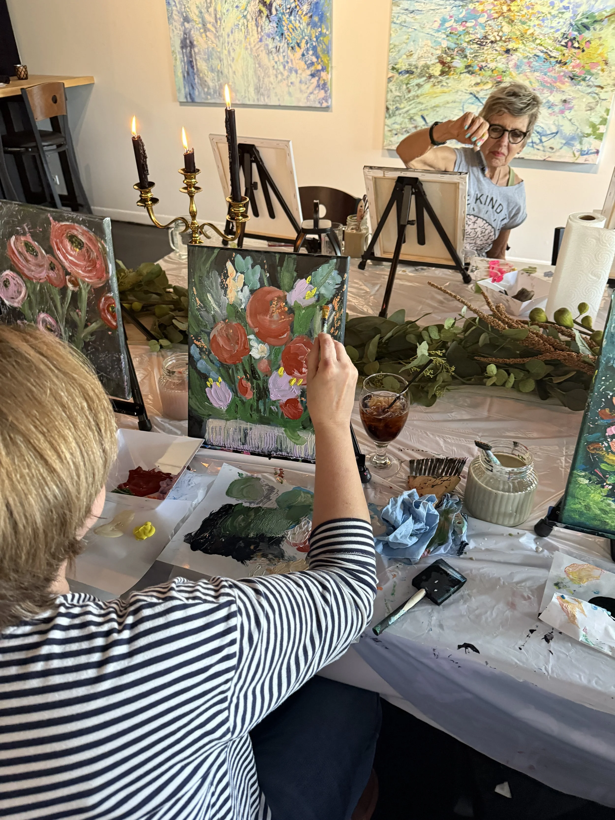 A woman with short gray hair and glasses, painting flower art on a canvas at an art studio table, surrounded by art supplies, with large colorful paintings on the walls behind her.
