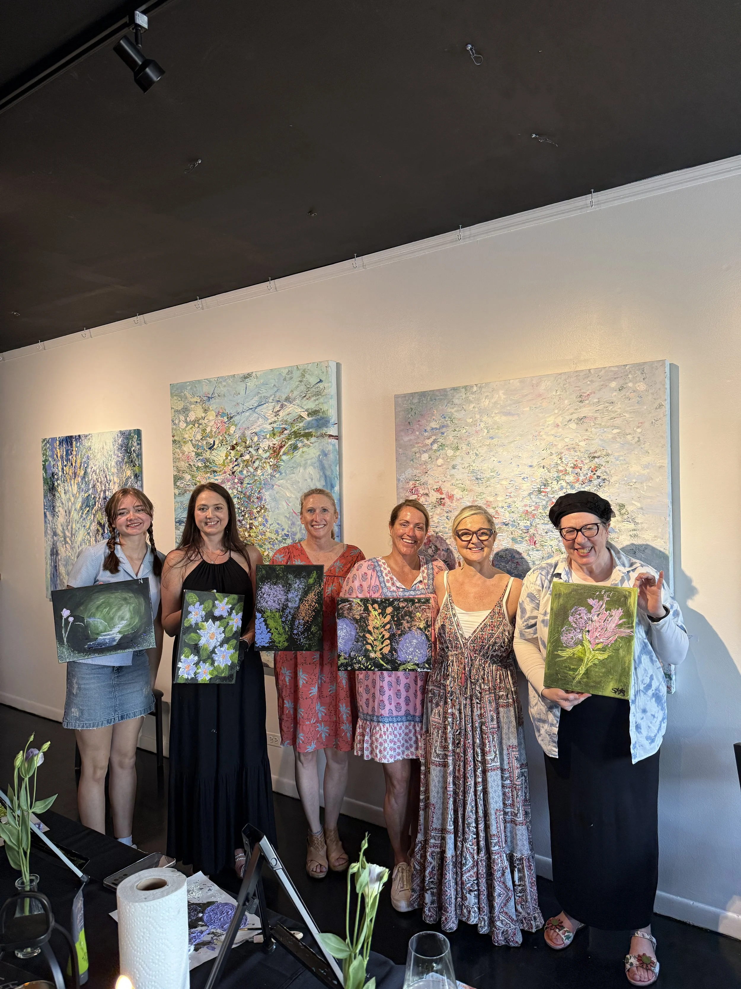 Six women standing together, smiling, holding paintings, in an art gallery with abstract artwork on the walls.