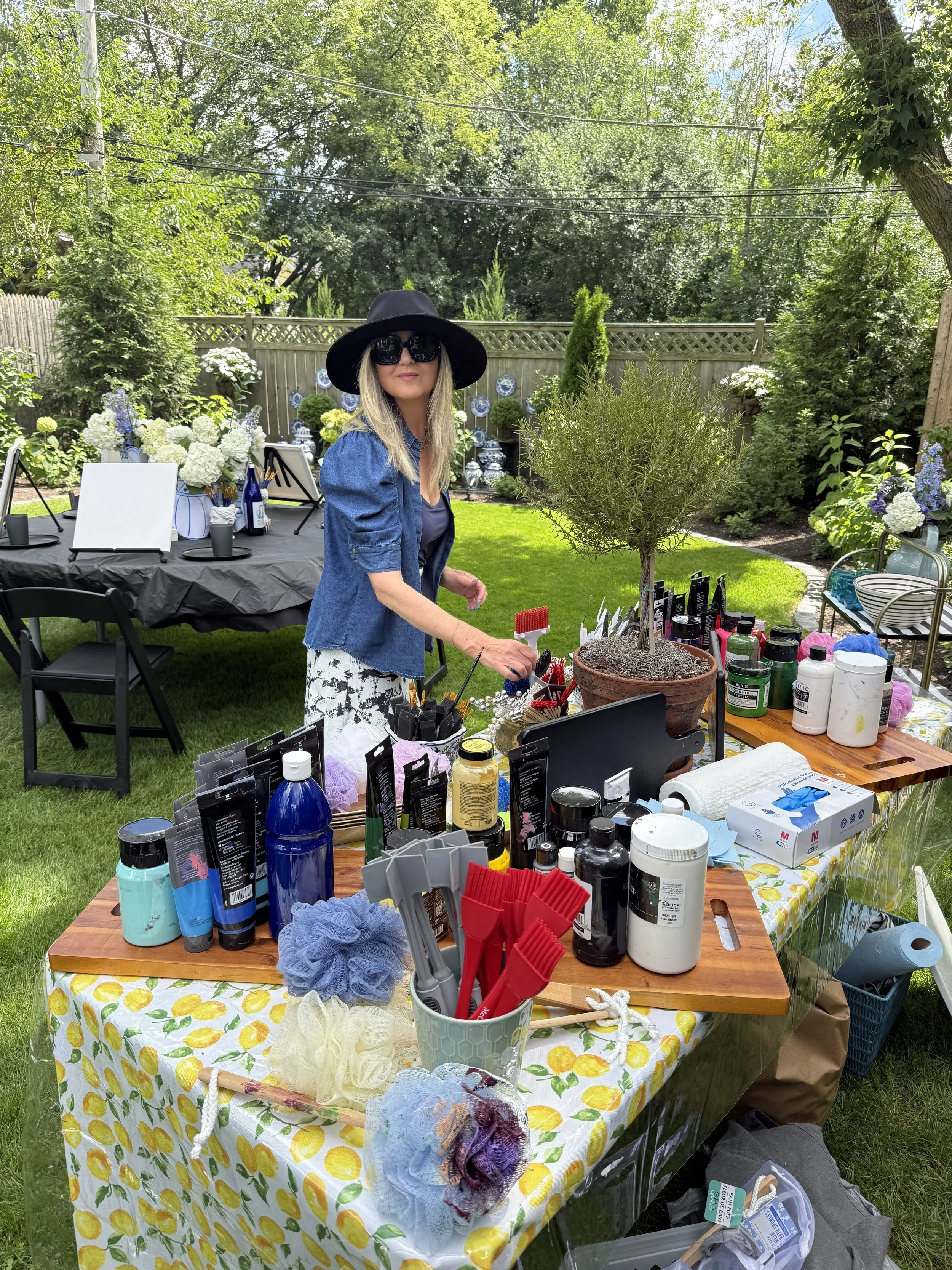A woman wearing a black wide-brimmed hat and large sunglasses stands near a table with various art supplies on a sunny backyard lawn. The table has a yellow tablecloth with a lemon pattern, and is filled with bottles, brushes, and other artistic item