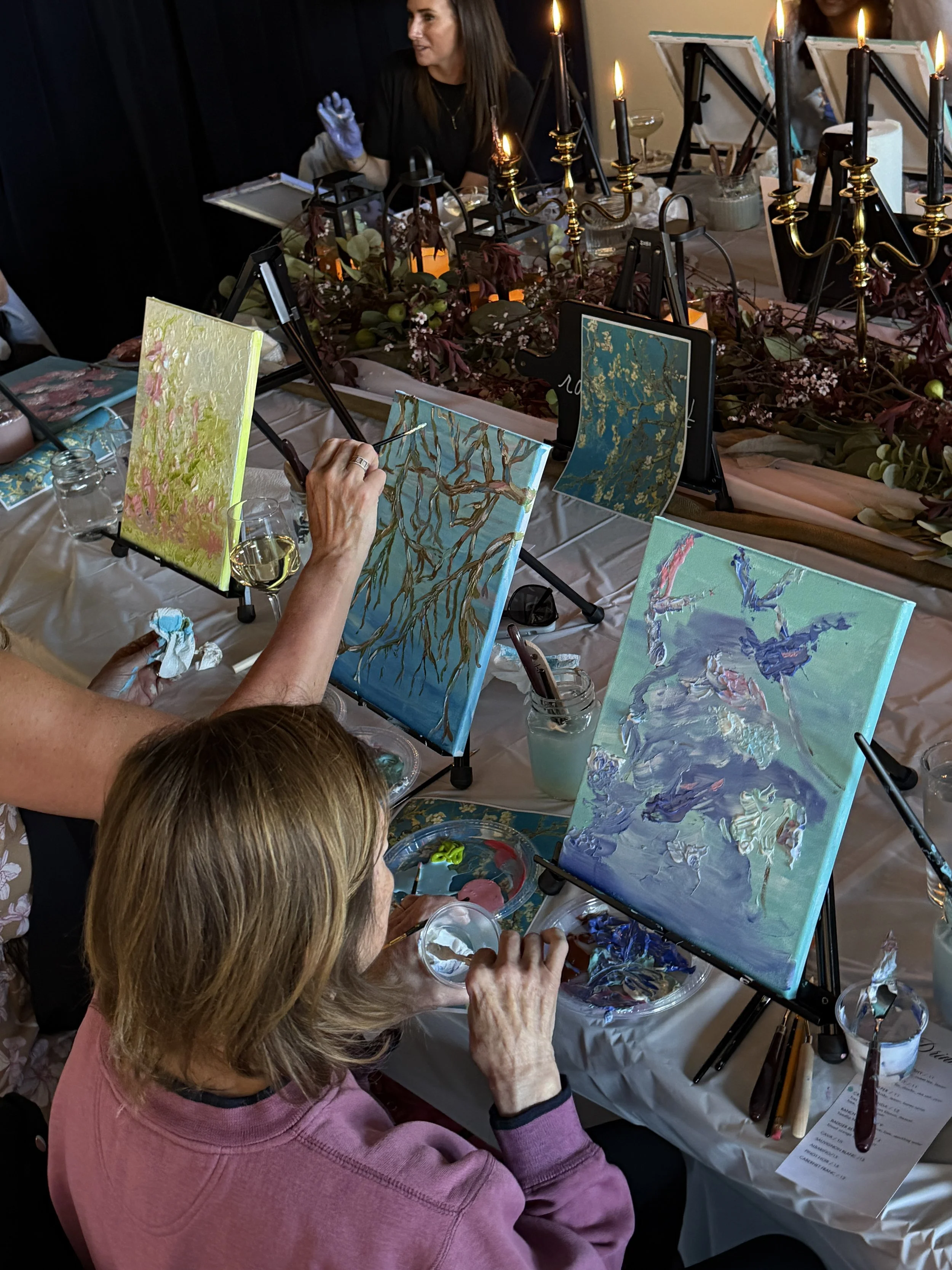 People painting at an art class with canvases, paints, and brushes, while a woman in a pink sweatshirt focuses on her artwork.