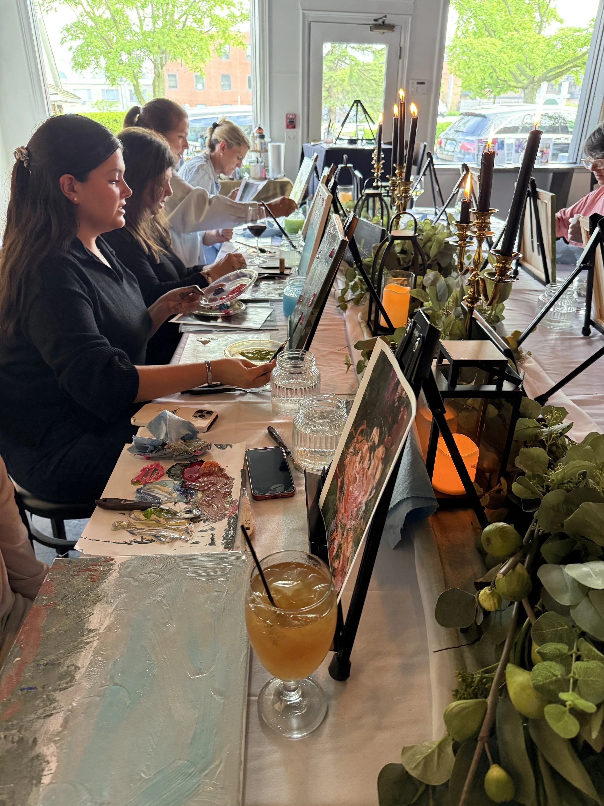 A group of people sitting at a long table during an art class or painting session, with art supplies and partially completed paintings in front of them. The table is decorated with candles and greenery. Large windows reveal trees and parked cars outs