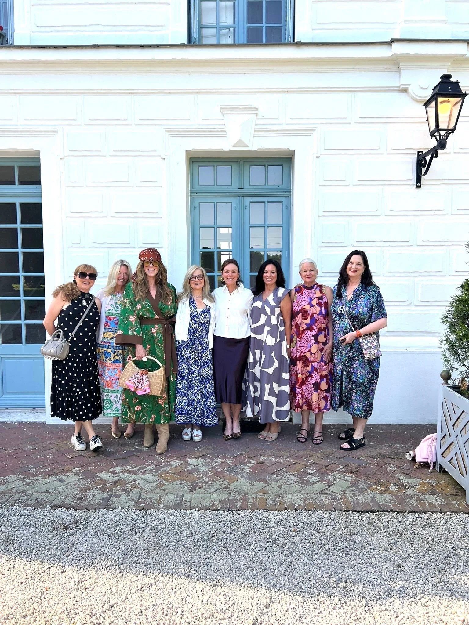 Group of eight women standing in front of a white building with blue door and window, smiling and posing for a photo.