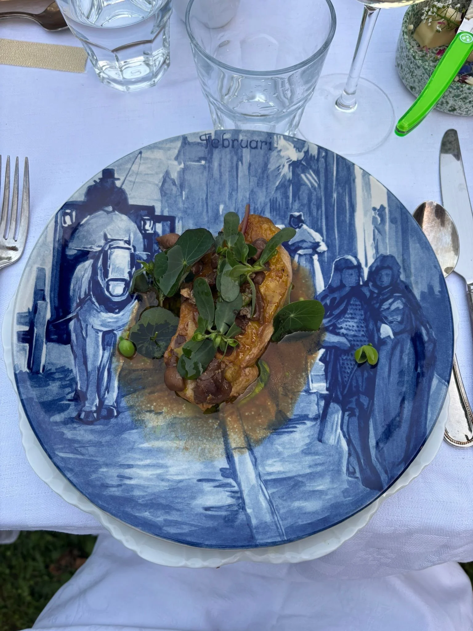 A gourmet dish served on a blue and white patterned plate, garnished with greens, with a fork on the left and a knife on the right, set on a white tablecloth alongside empty glasses and a glass of water.