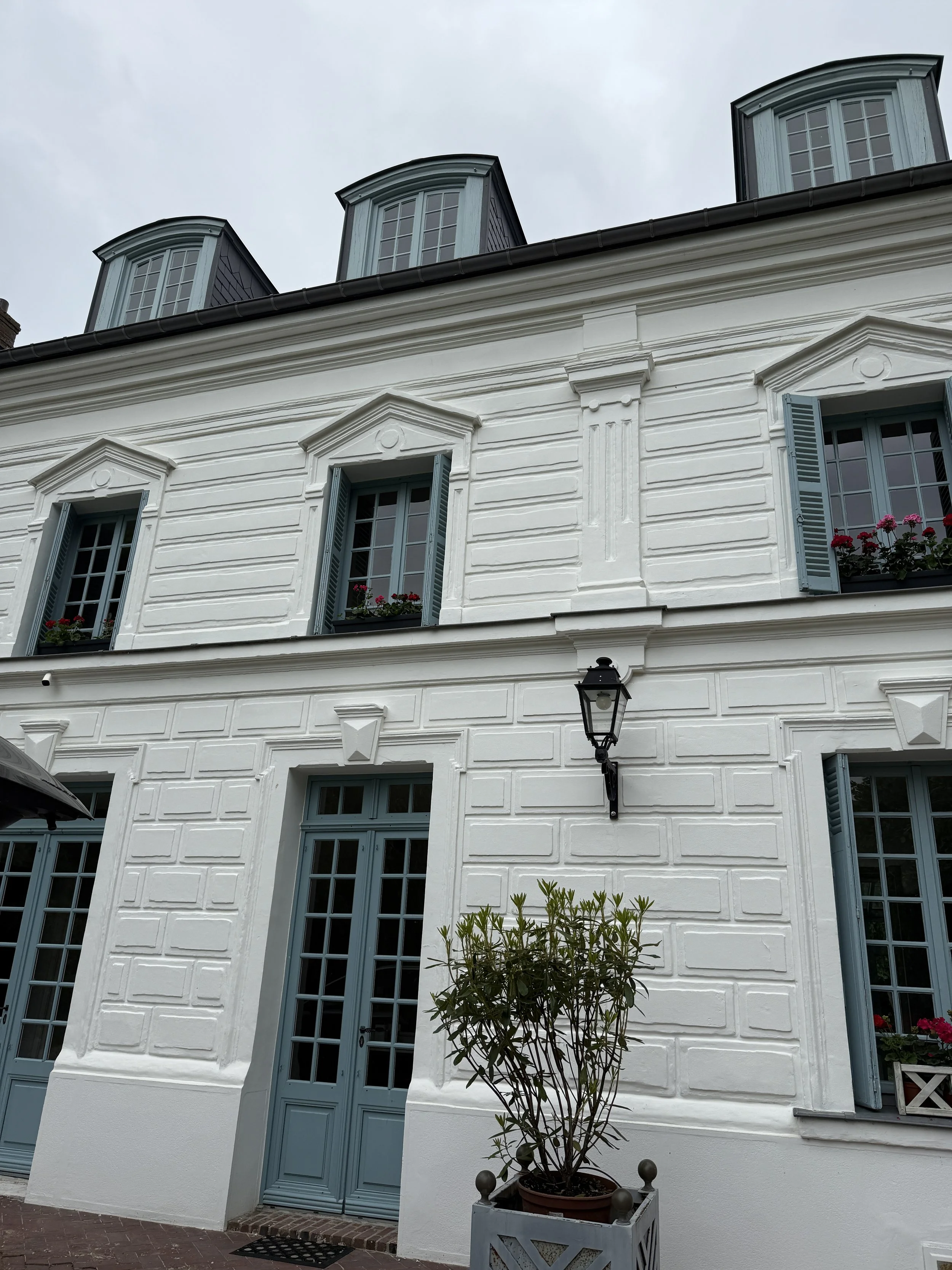 White building with blue shutters, multiple windows, and dormer windows on the roof. There is a black street lamp and a potted plant in front.