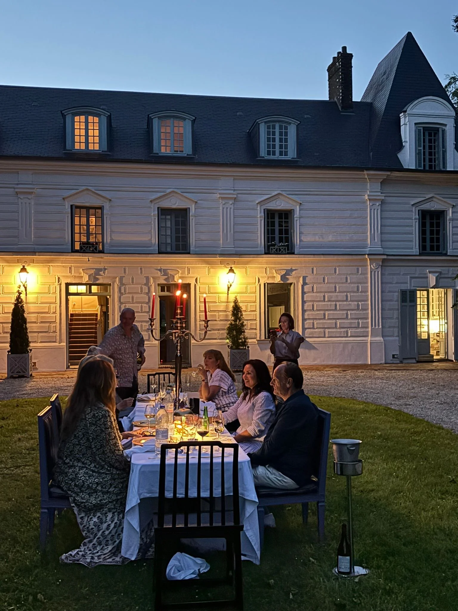 People dining outdoors at twilight in front of a white historical building with lit windows and a gray roof, with some candles and a chandelier on the table.