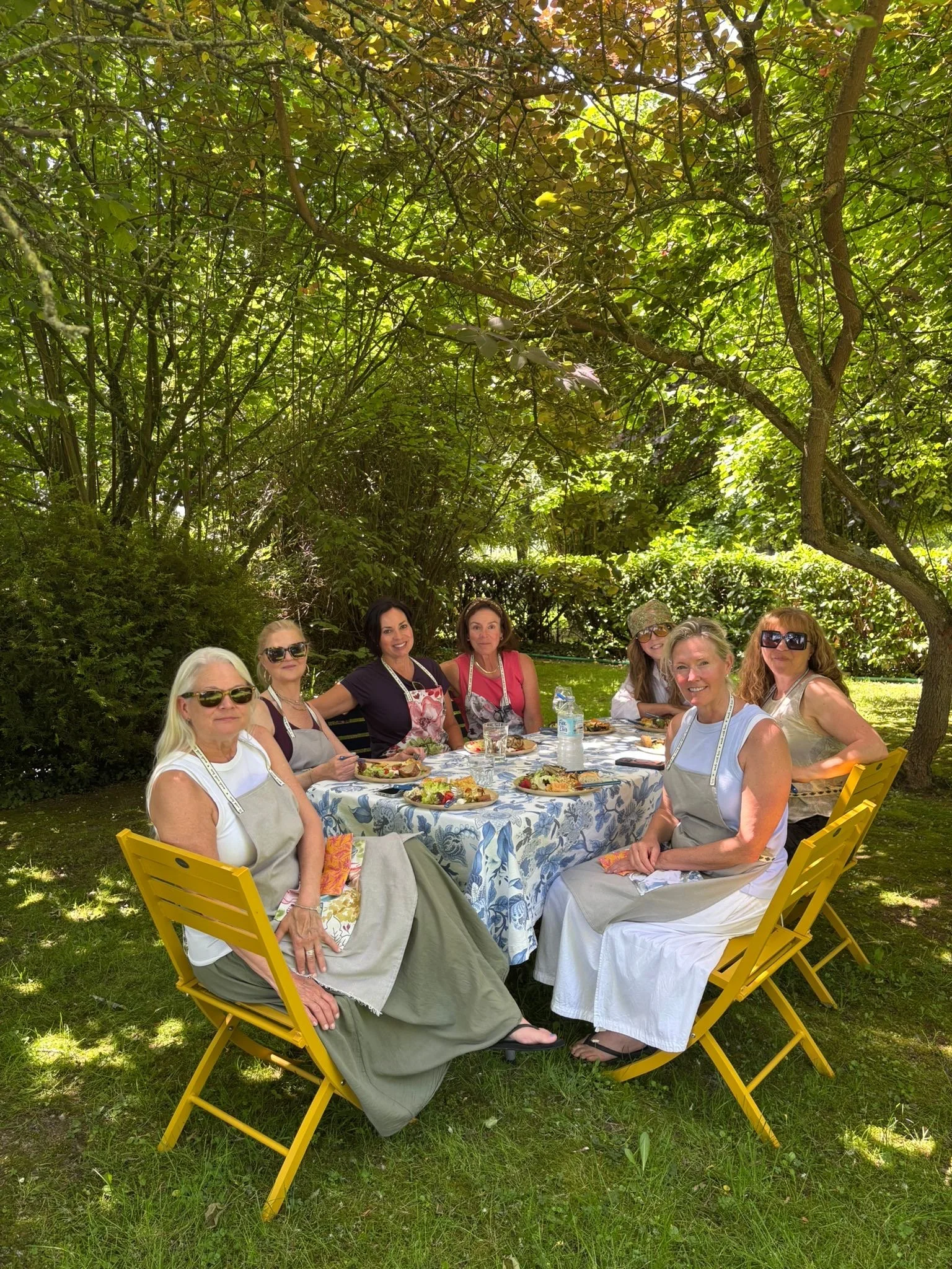 A group of women sitting around a table outdoors under a large tree, enjoying a meal together on a sunny day.