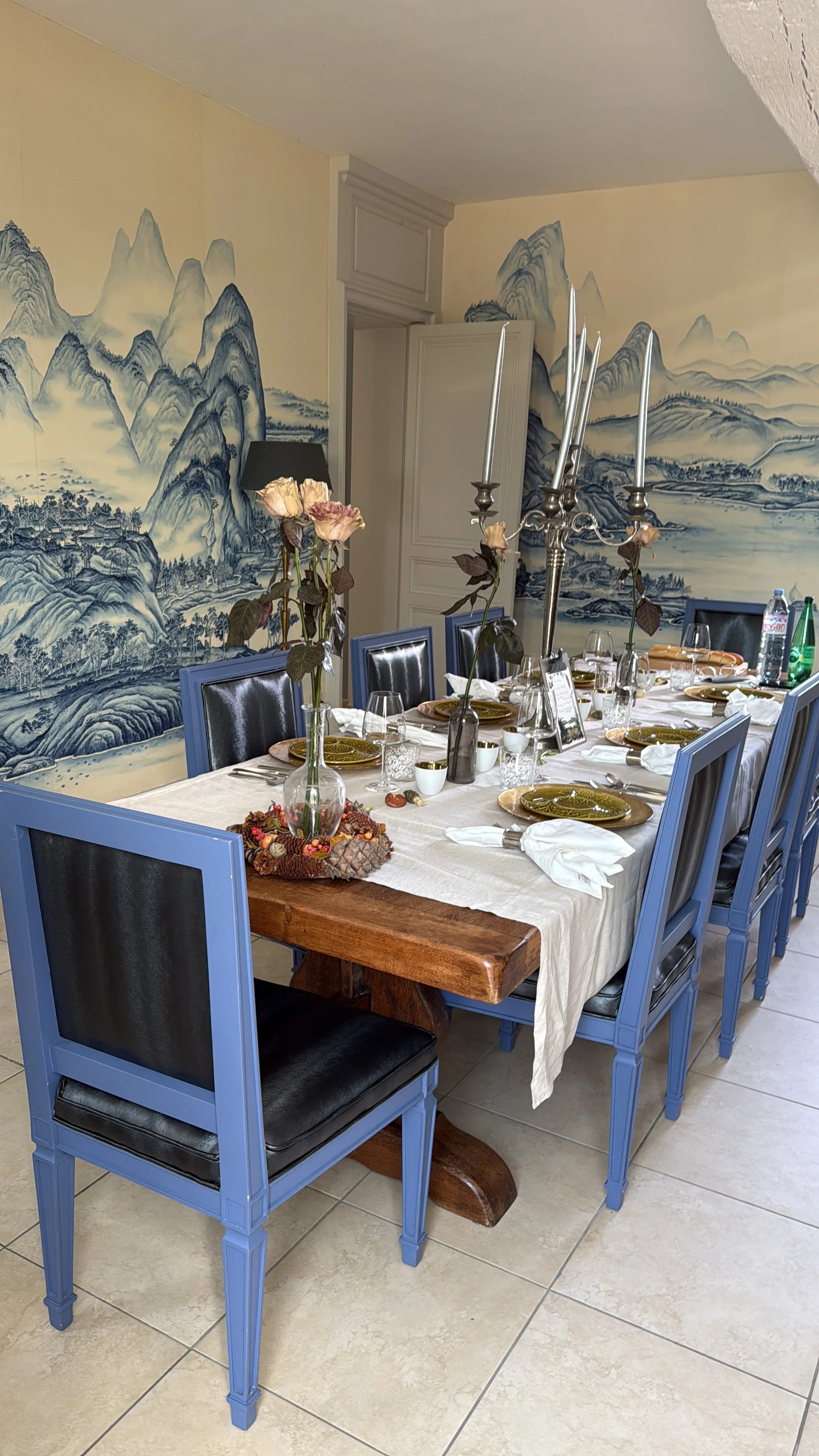 A dining table set for a meal with plates, glasses, and silverware, decorated with tall vases of dried roses and a large silver candelabrum, in a room with a mural of traditional Chinese landscape paintings on the wall.