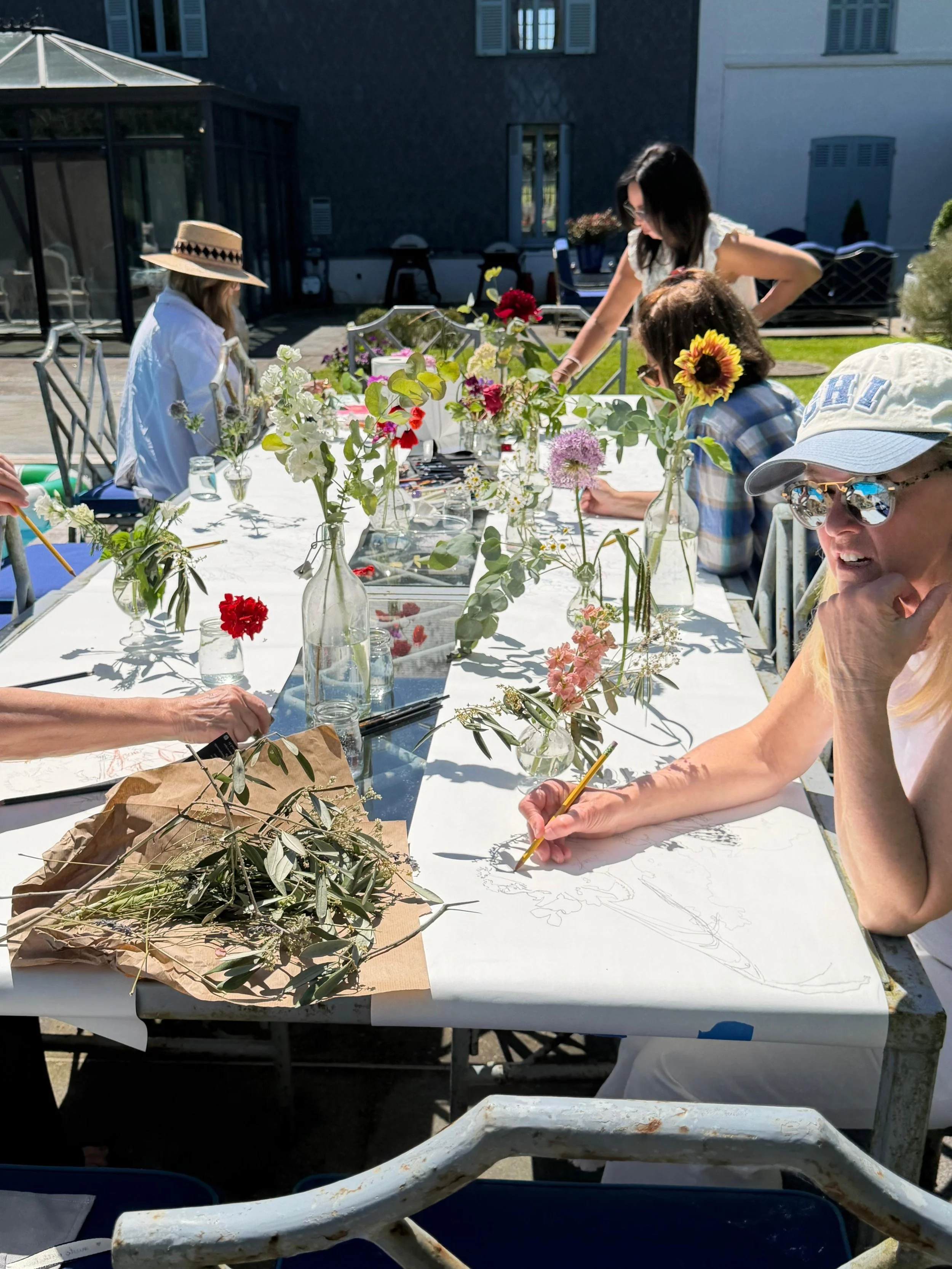 Group of women creating flower arrangements on a long outdoor table, with various vases and flowers, in a sunny backyard.