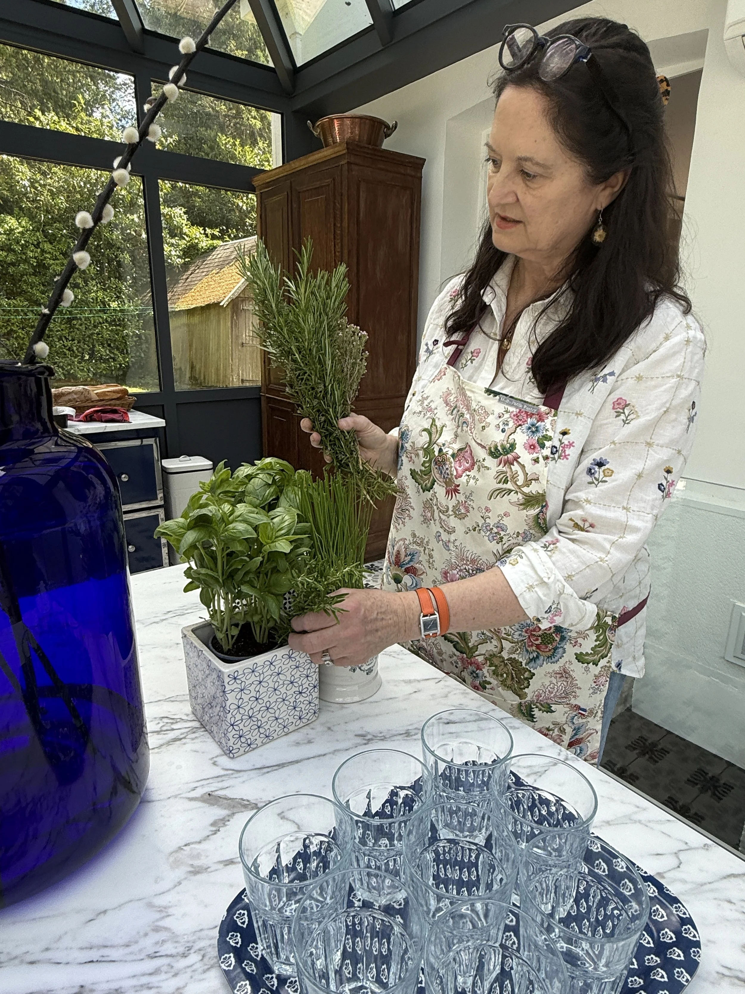 A woman with glasses on her head and wearing a floral apron is holding a potted herb plant with green leaves. She is standing at a marble table with multiple empty glasses arranged on a dark blue patterned placemat. The background shows a sunlit cons