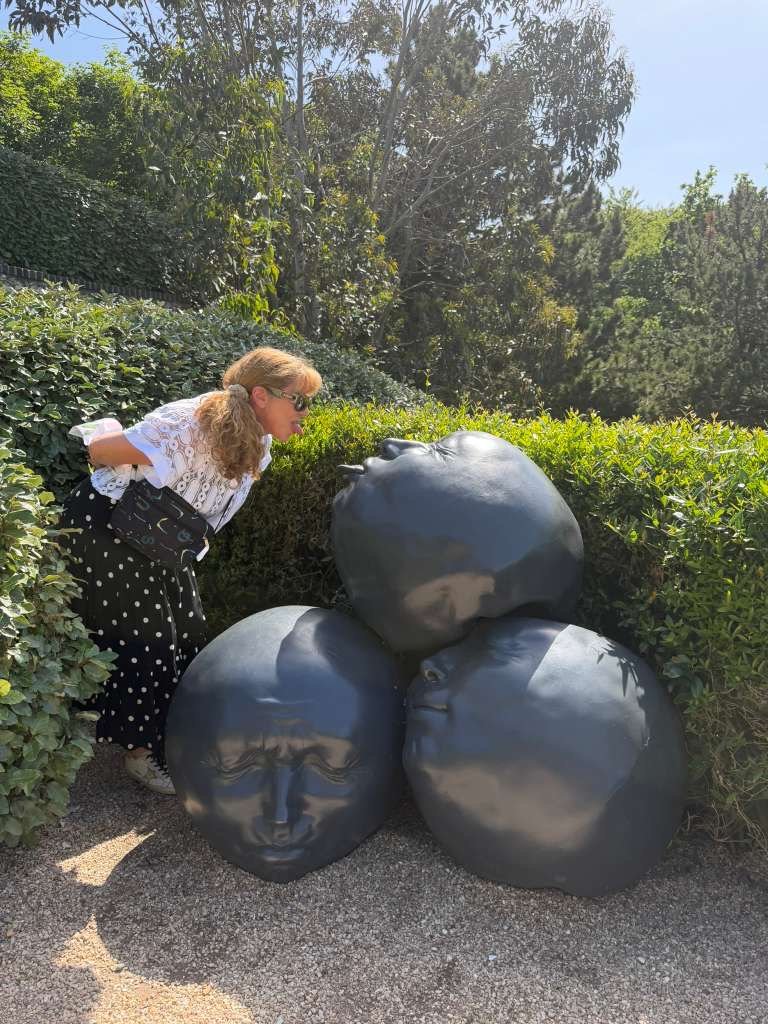 A woman with curly blonde hair, wearing sunglasses, a white blouse, and a black skirt with white polka dots, leans in to observe four black stone sculptures of human faces outdoors on a sunny day, surrounded by green bushes and trees.
