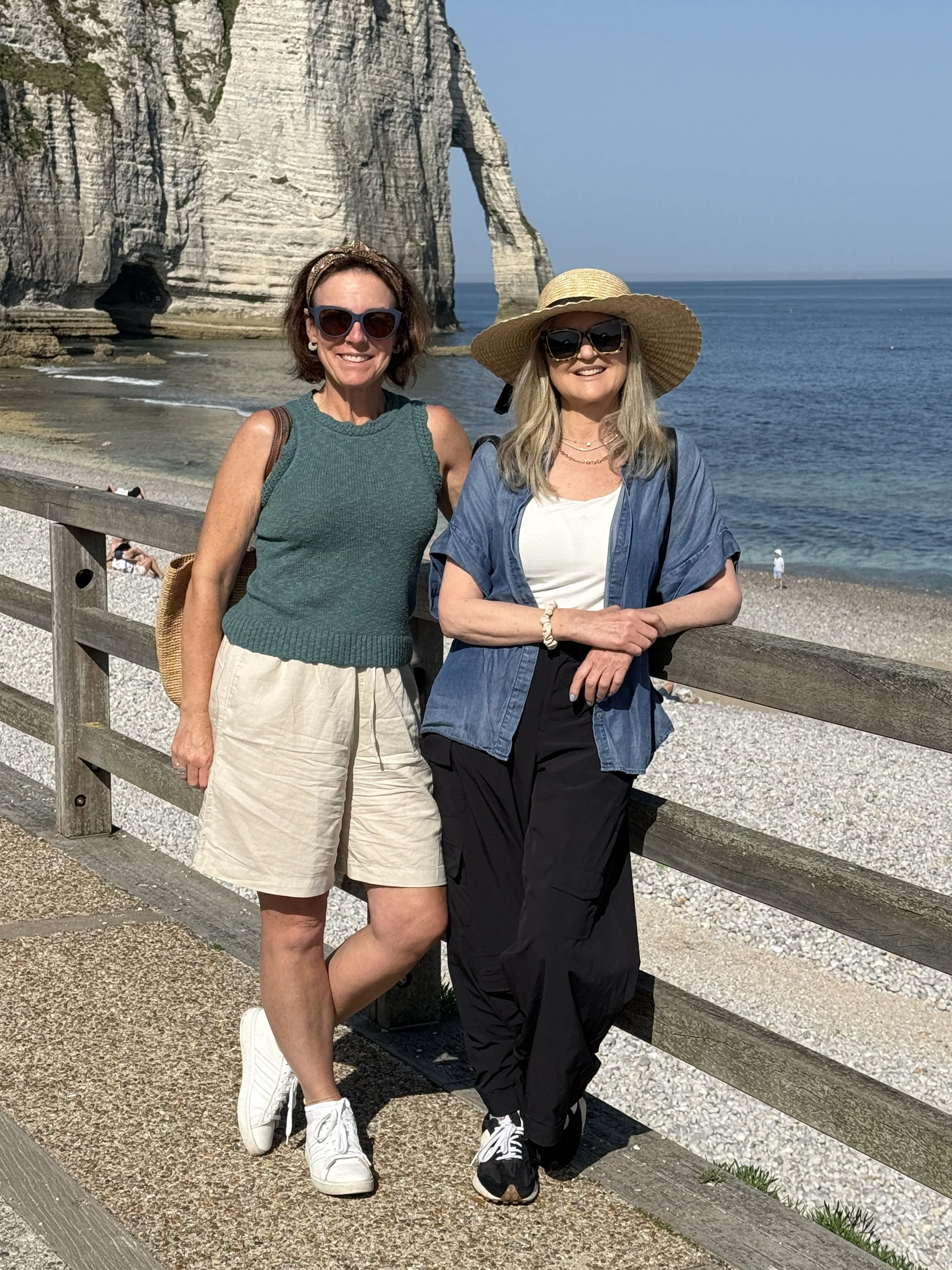 Two women standing on a seaside promenade with large rock formations and ocean in the background. One is wearing a teal sleeveless top, beige shorts, white sneakers, and sunglasses. The other is wearing a wide-brimmed straw hat, a denim shirt over a 