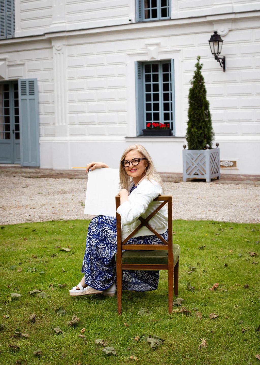 A woman with glasses and blonde hair sitting on a wooden chair outdoors, smiling at the camera, with a white building and green lawn in the background.
