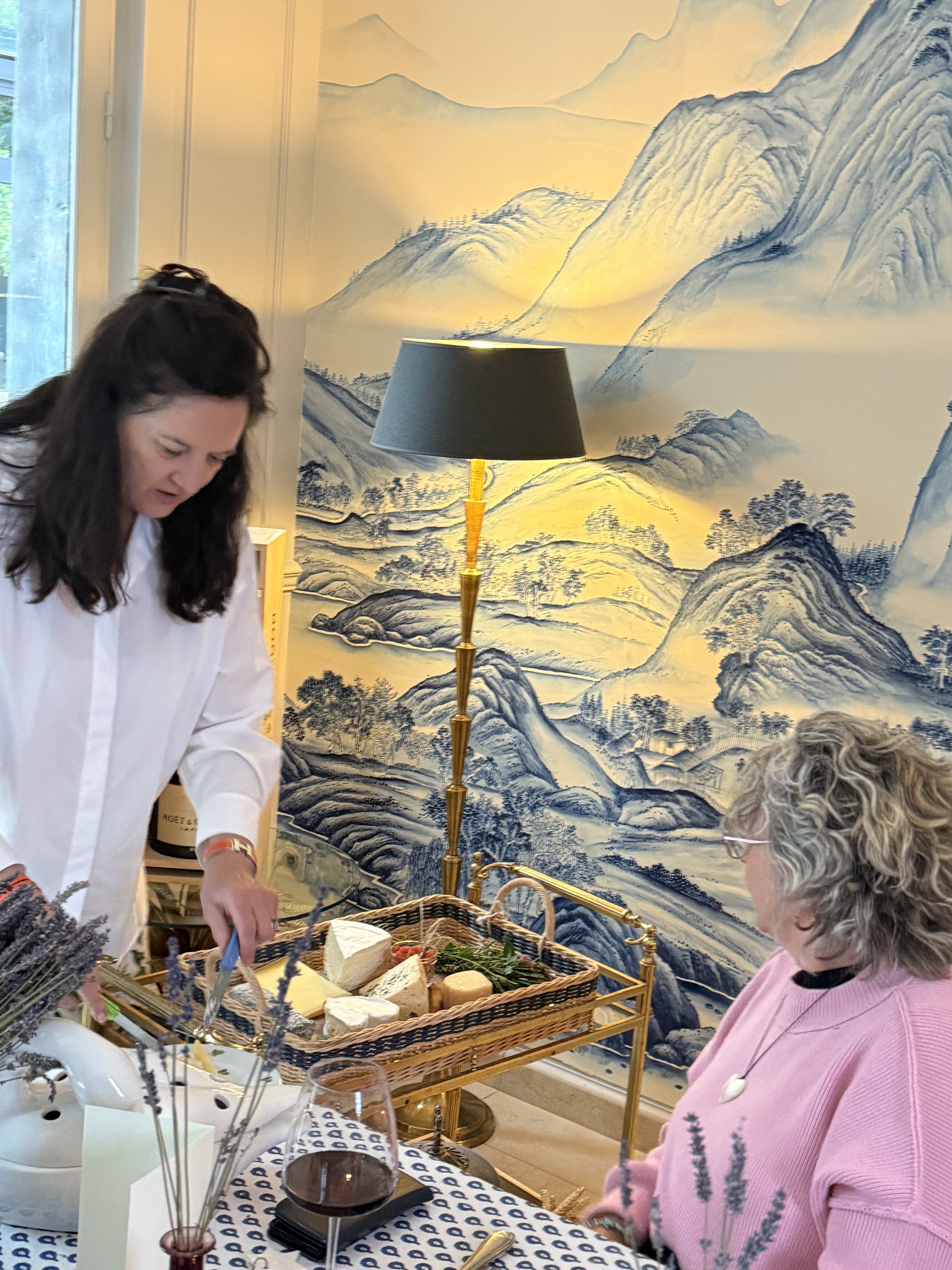 Two women are at a table with food and wine glasses, with a blue and white patterned tablecloth; one woman in a white shirt is reaching towards the basket of bread, the other woman in a pink sweater is observing; a black and gold floor lamp is in the
