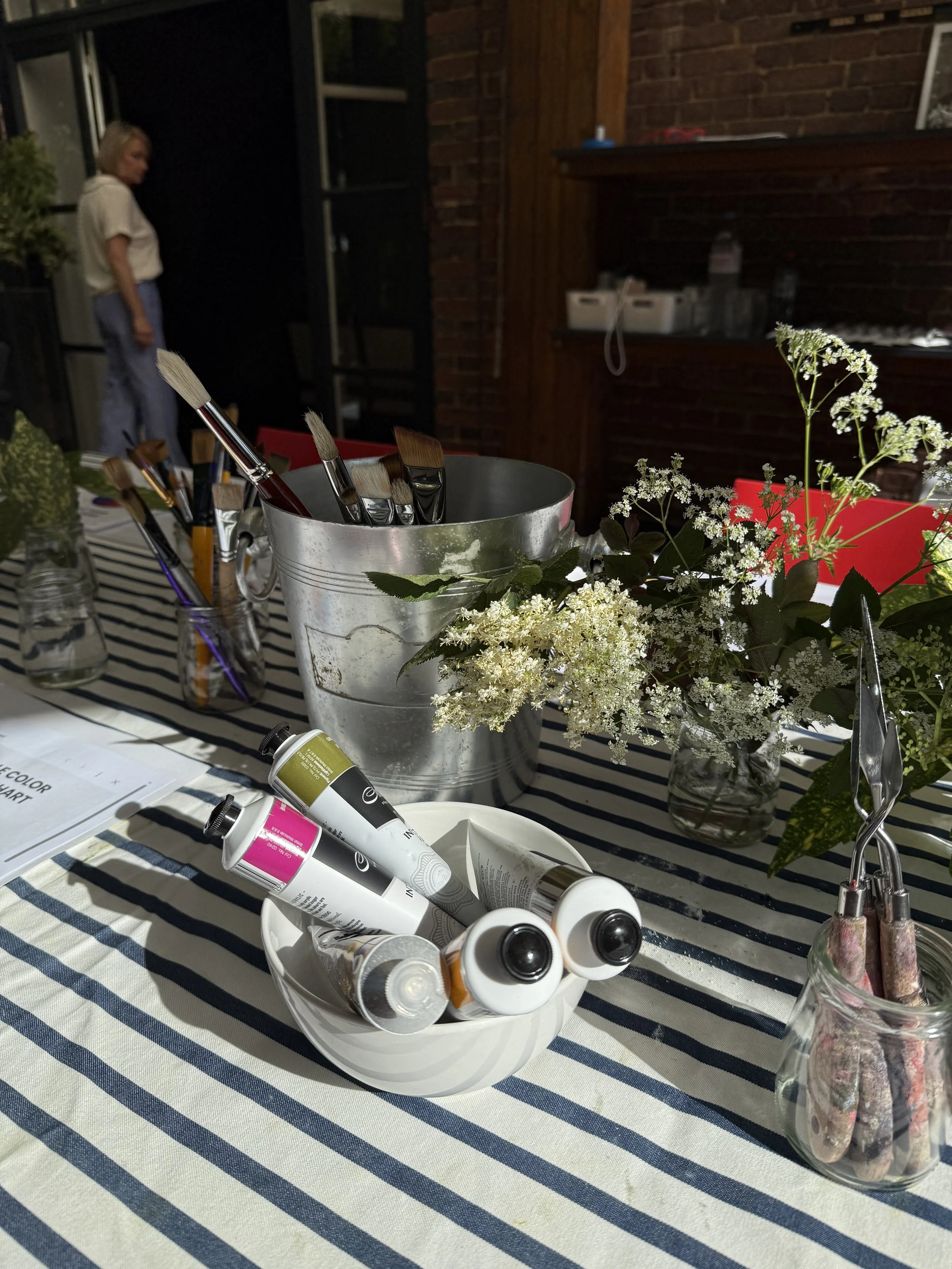 A table decorated with flowers and art supplies, including paint tubes and brushes, with a woman standing in the background of an outdoor patio at night.