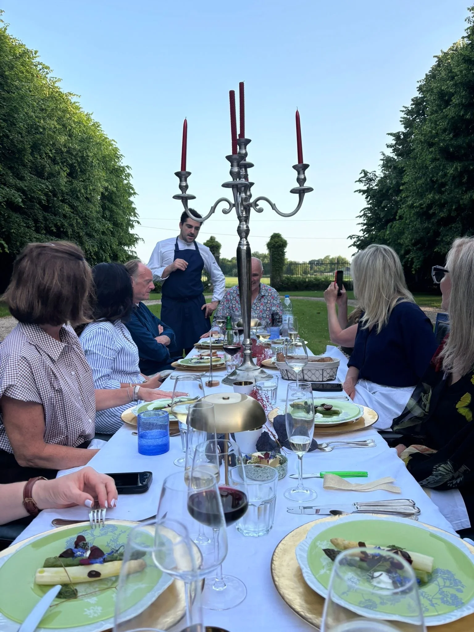 A group of people sitting at a long outdoor table for a meal, with a man in a white shirt and apron standing and talking, and a large candelabra with three red candles as a centerpiece.