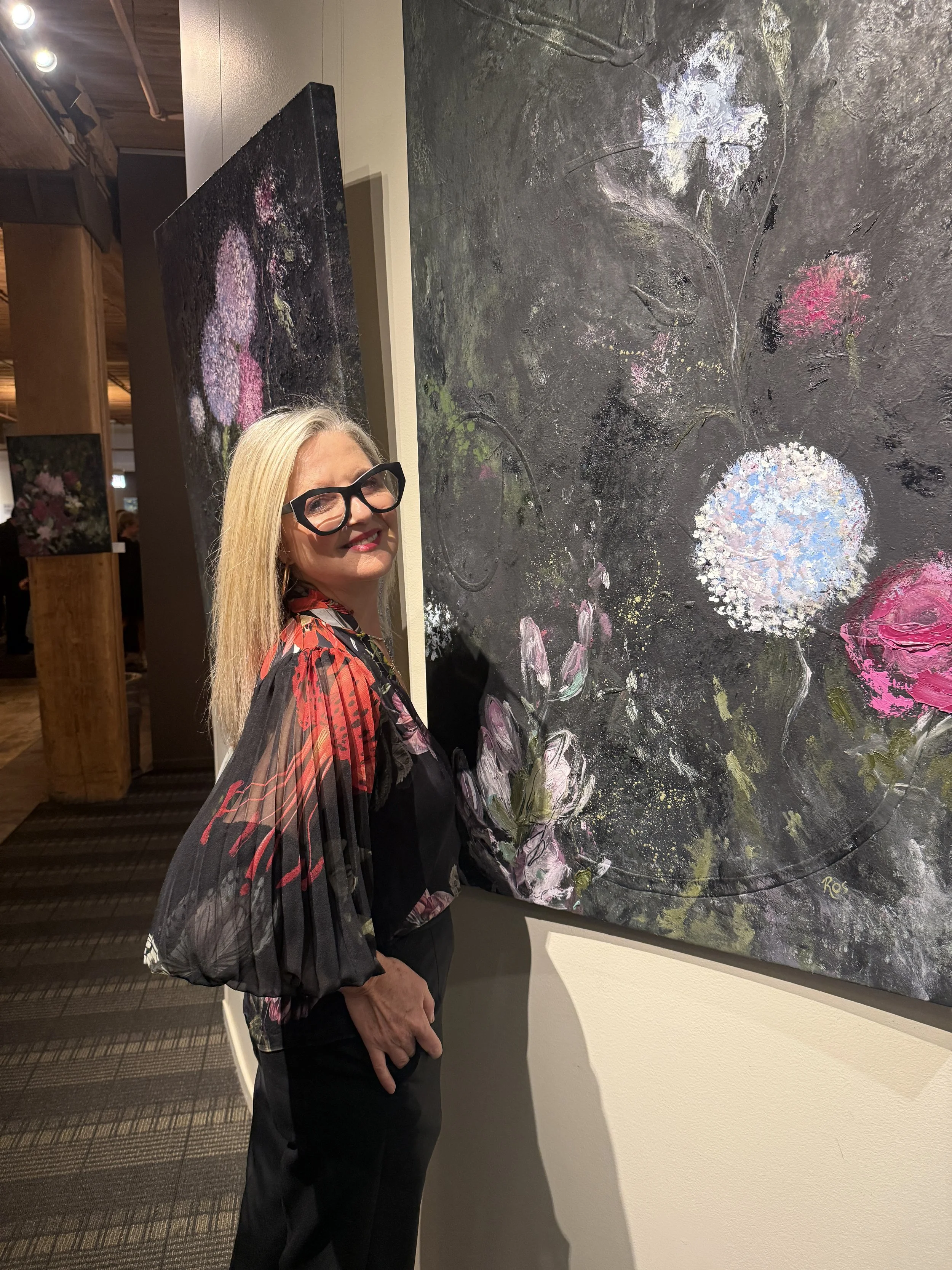 A woman with long blonde hair wearing glasses and a black dress with a sheer, colorful floral-patterned cape is standing in front of a large abstract floral painting at an art gallery.