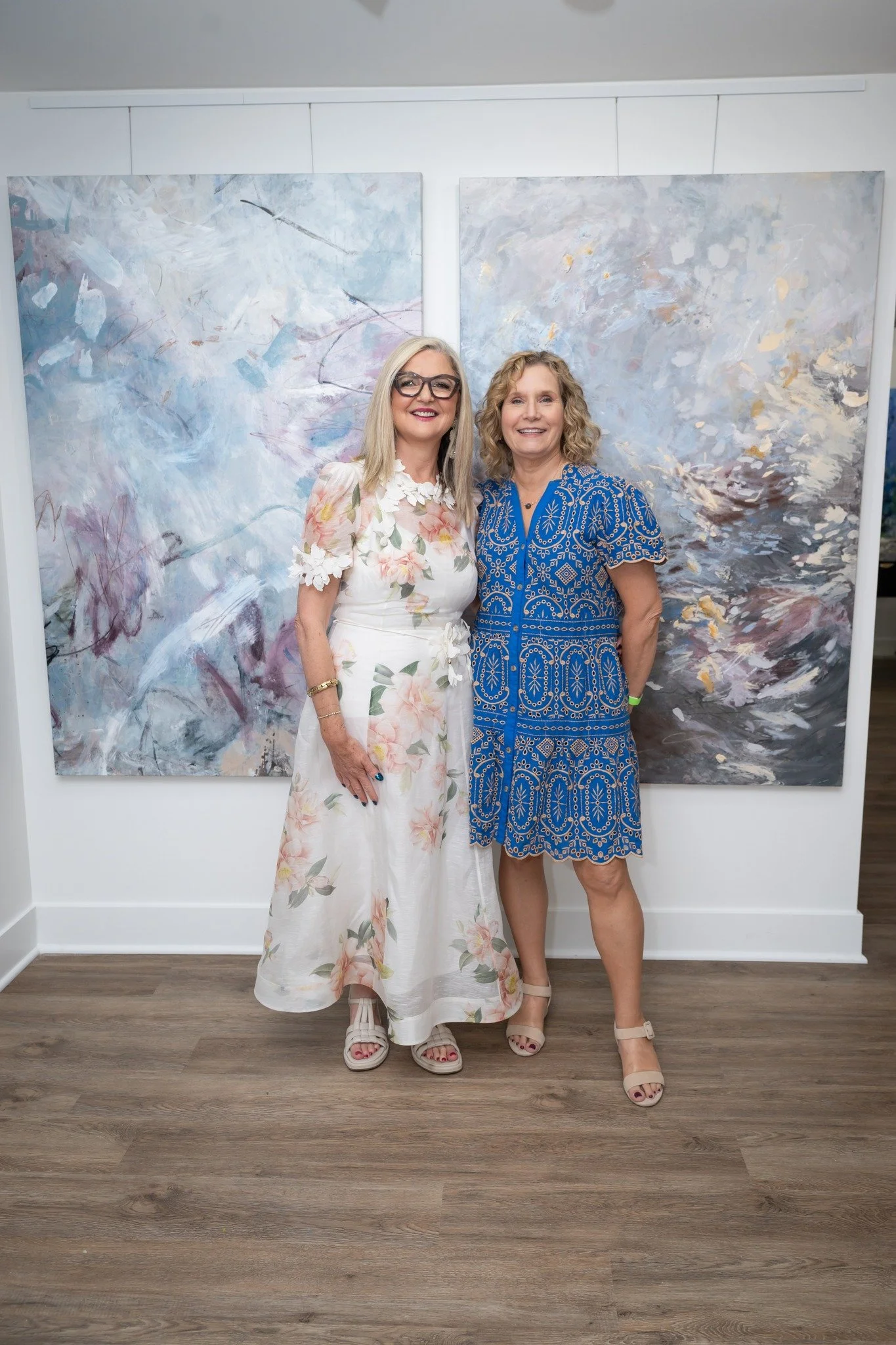 Two women stand side by side, smiling, in front of two large abstract paintings. The woman on the left wears a white floral dress with pink and green accents, and beige sandals. The woman on the right wears a blue dress with white geometric patterns and nude high heels. The setting appears to be an art gallery with a wooden floor.