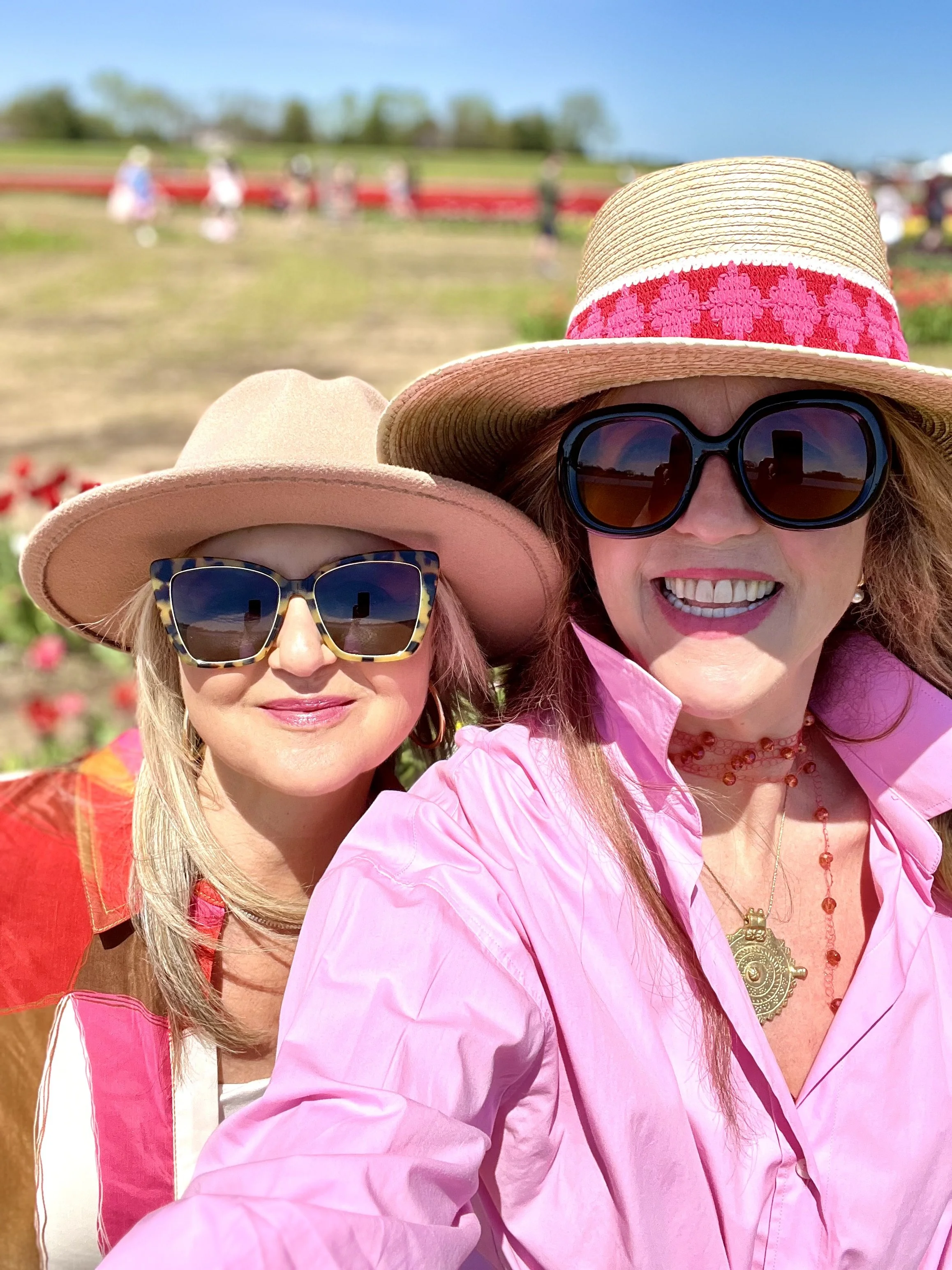 Two women wearing sunglasses and sun hats taking a selfie outdoors at a flower field on a sunny day.