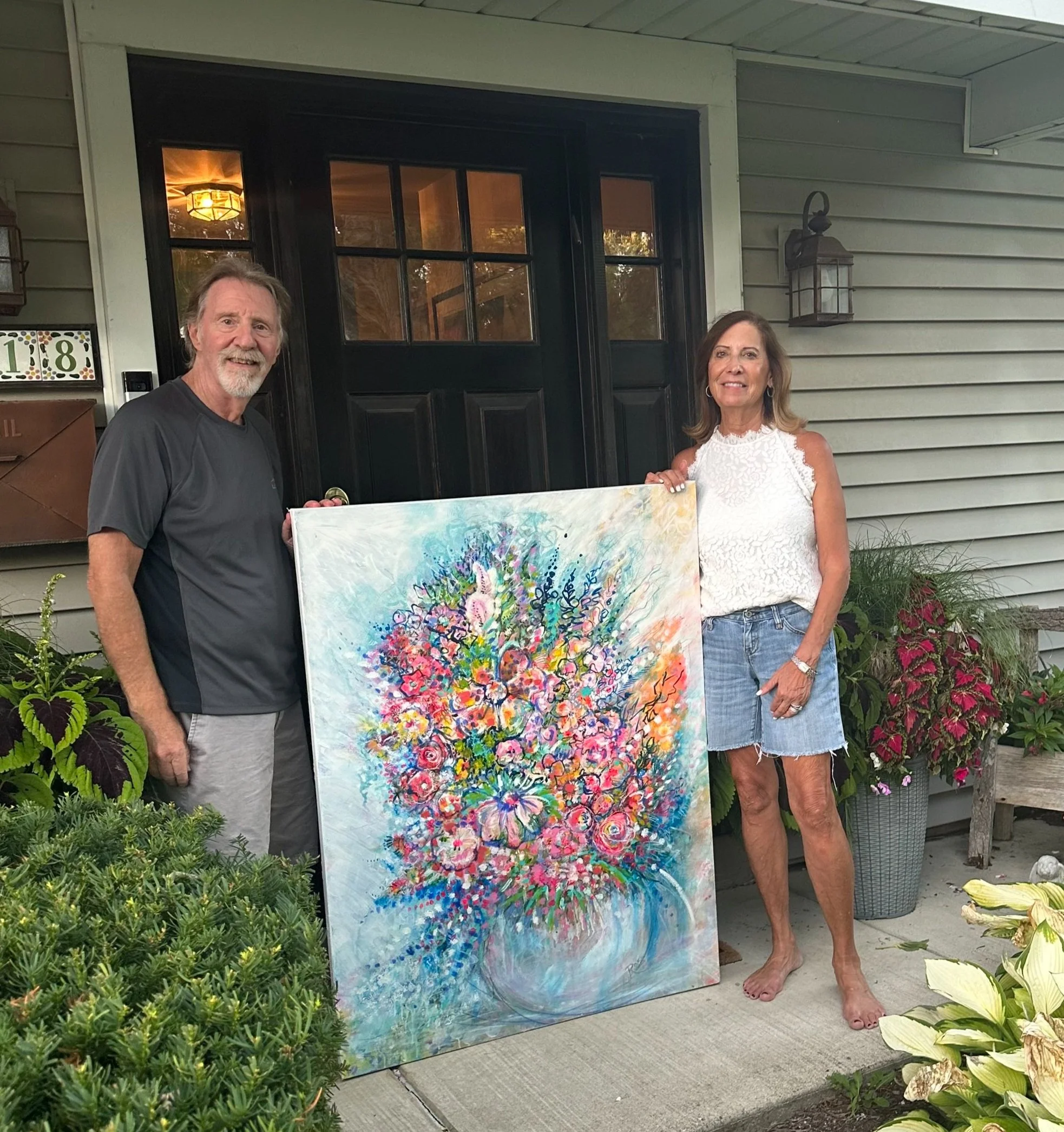 A man and woman standing outdoors in front of a house holding a colorful abstract painting. The man is wearing a dark T-shirt, has a beard, and gray hair. The woman is wearing a white lace top, has shoulder-length brown hair, and is barefoot. There are green plants and flowers on either side of them, with a black door and house exterior in the background.